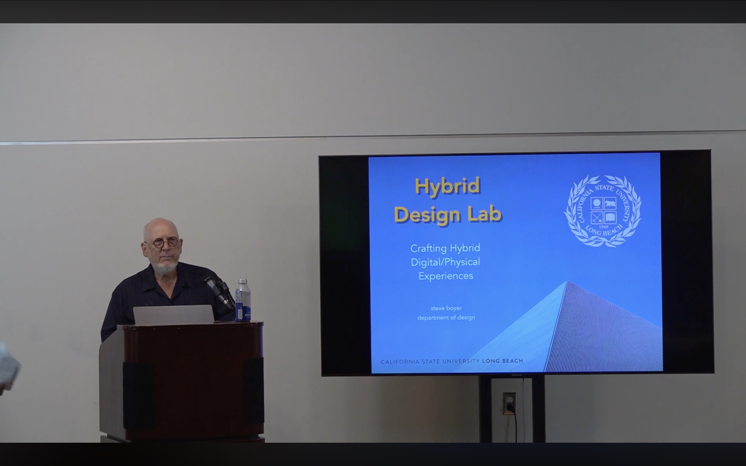 Steve Boyer behind a wooden podium and in front of a projector for a presentation on the Hybrid Design Lab