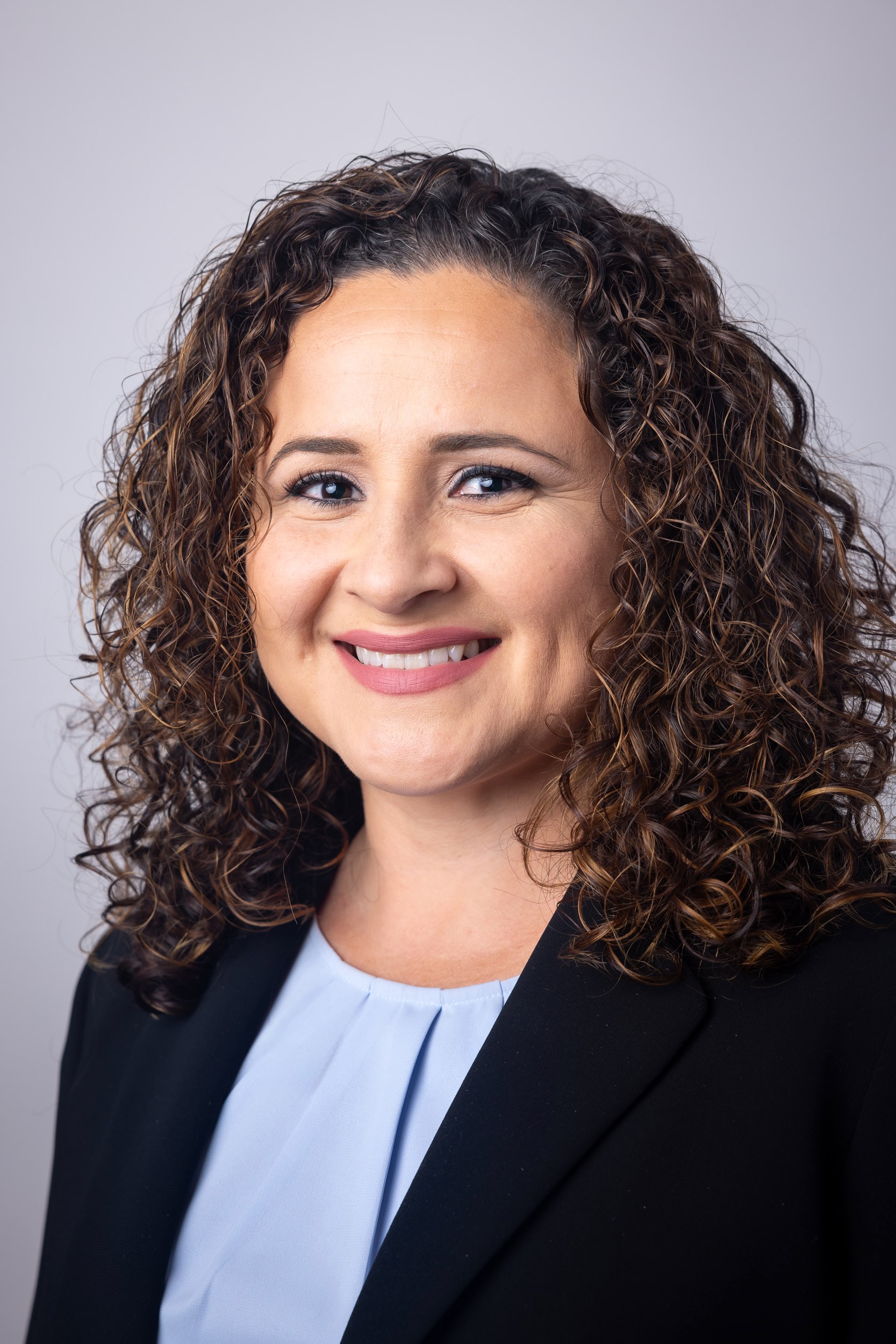 Professional headshot of Dr. Daisy D. Alfaro, a Latina with shoulder length curly hair, smiling, wearing a black blazer over a light blue blouse, against a neutral background.