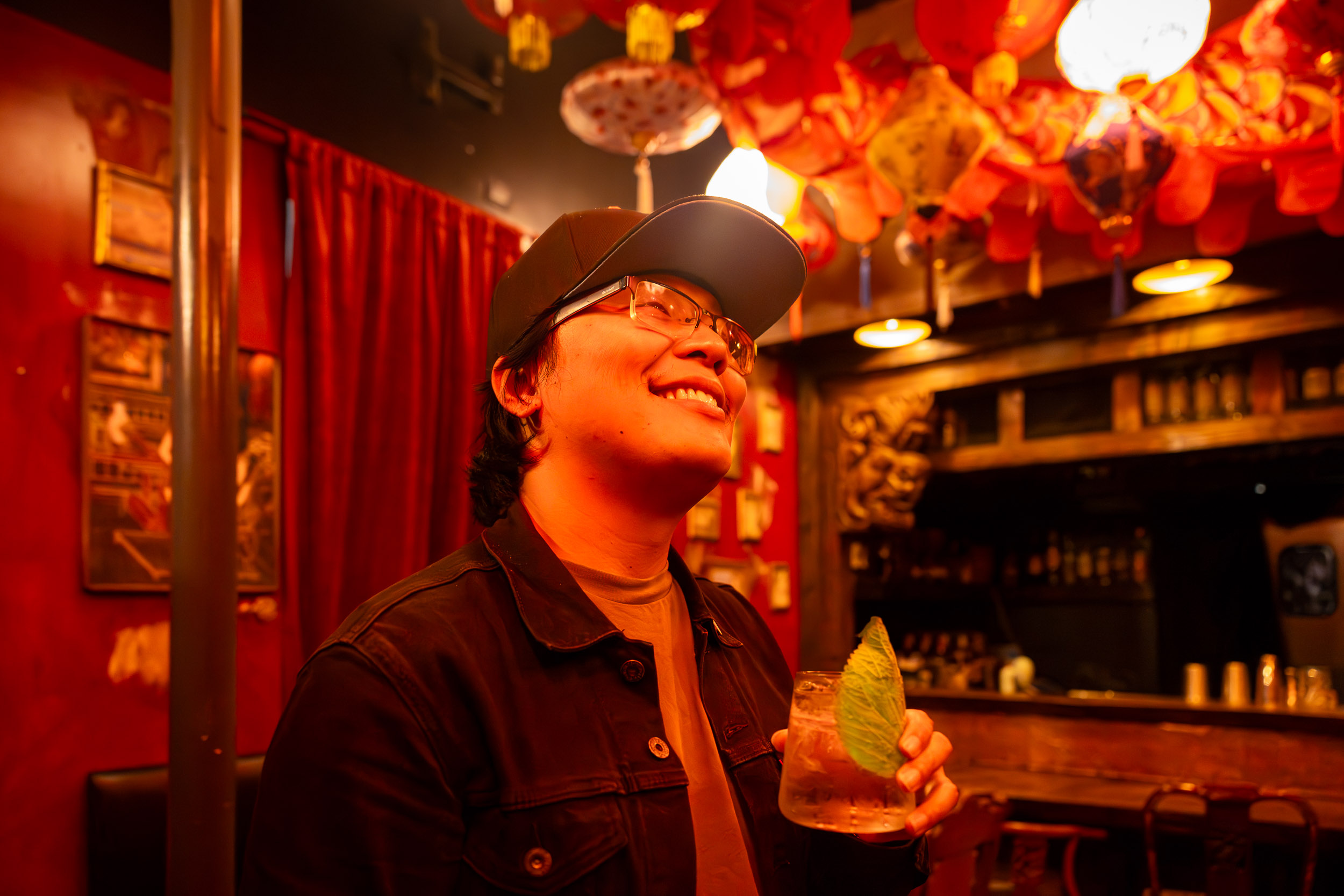 Person wearing a cap smiles while holding a drink in a dimly lit bar.
