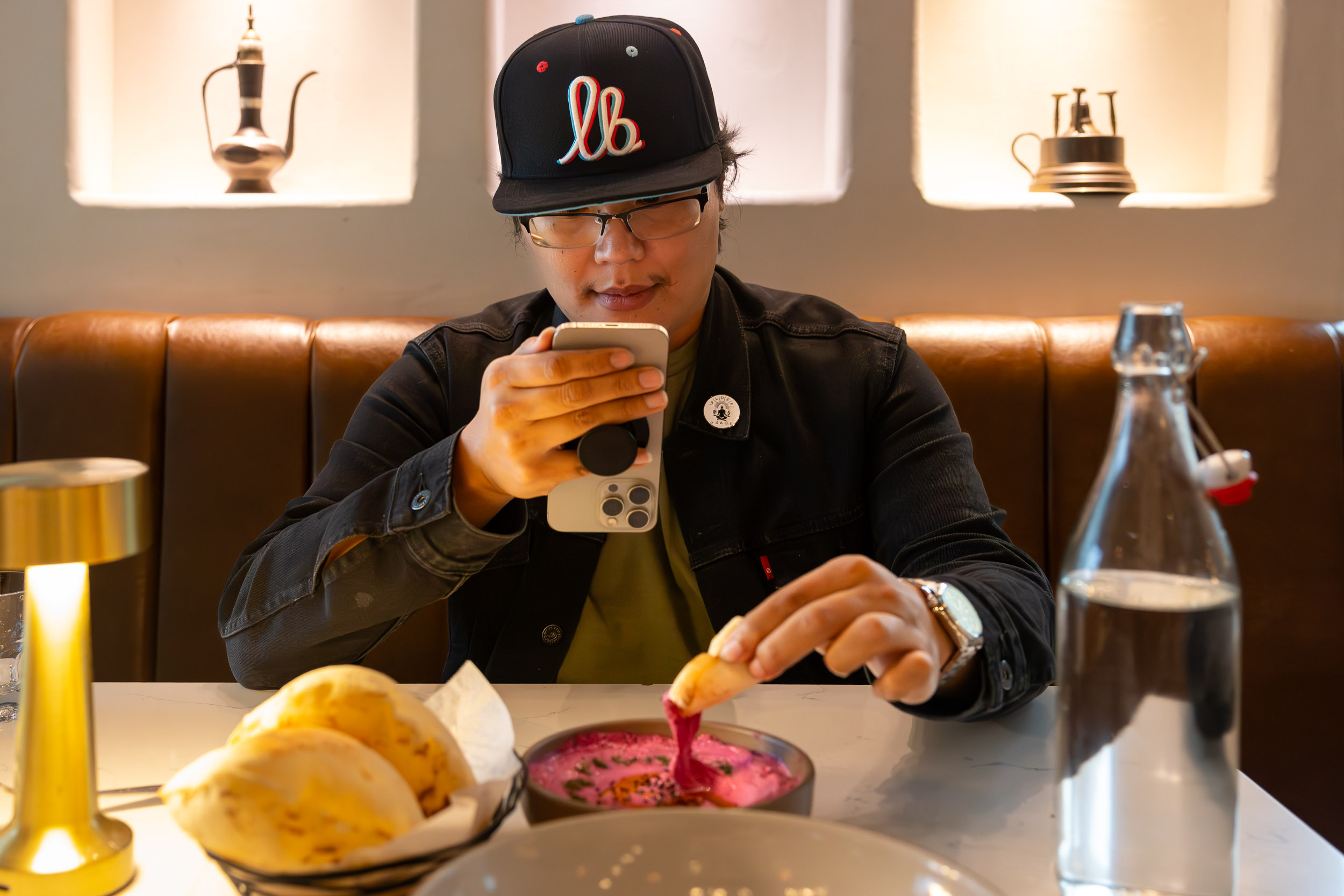 Person sits at a restaurant table taking a phone photo while dipping bread into a bowl of food.