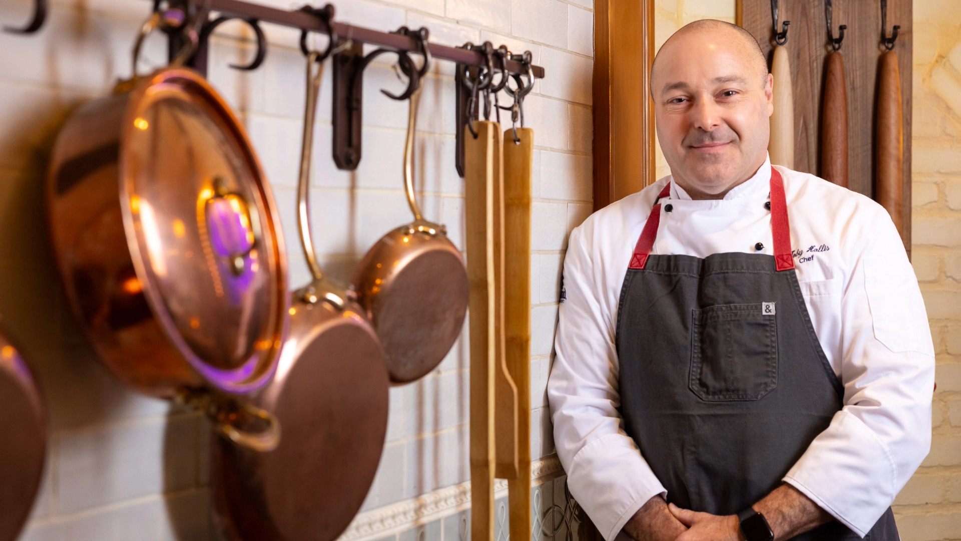 Toby Hollis stands beside a wall with copper pots and wooden rolling pins hanging.