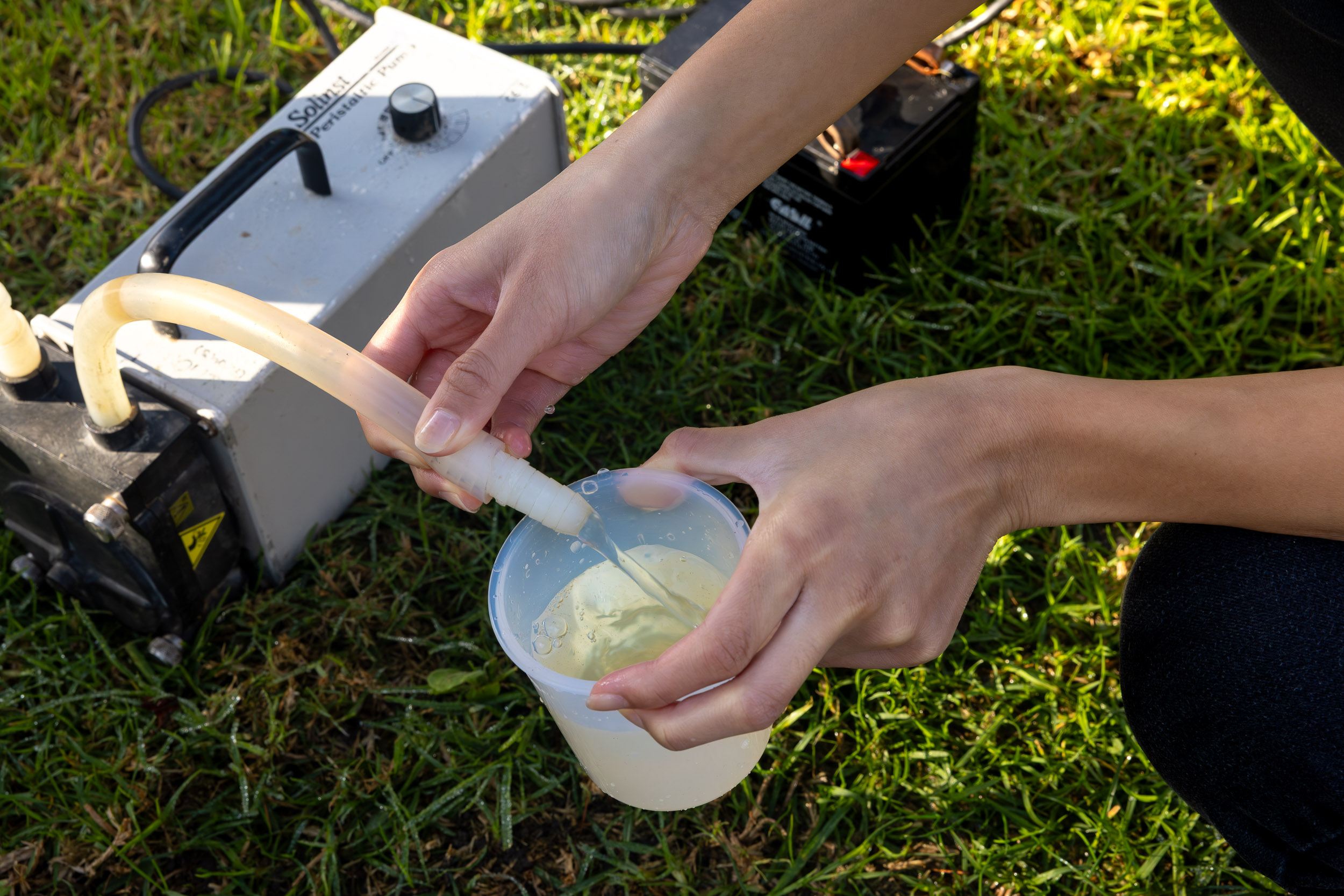 Hands hold a hose as liquid flows into a clear container during field sampling.