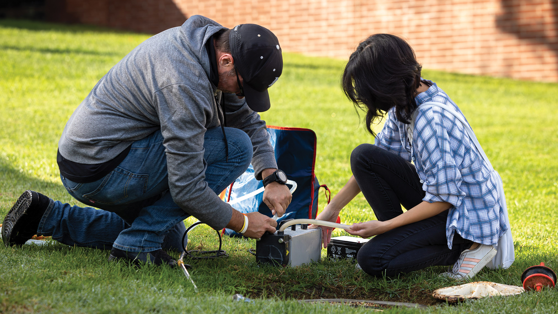 Two people kneel on a campus lawn operating scientific equipment while one person collects a liquid sample into a marked tube.