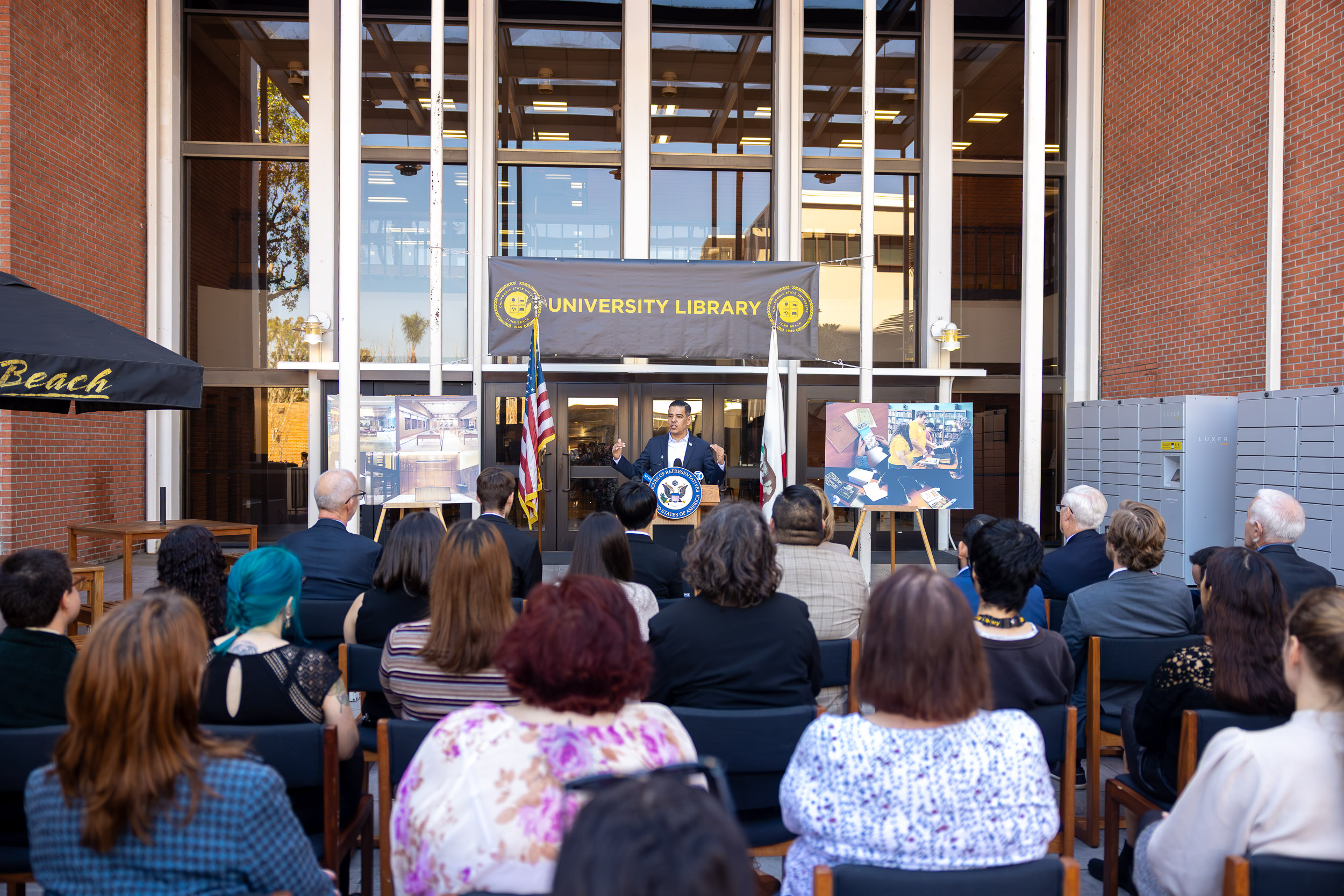 A person speaks at a podium outside the University Library as an audience sits facing the entrance.