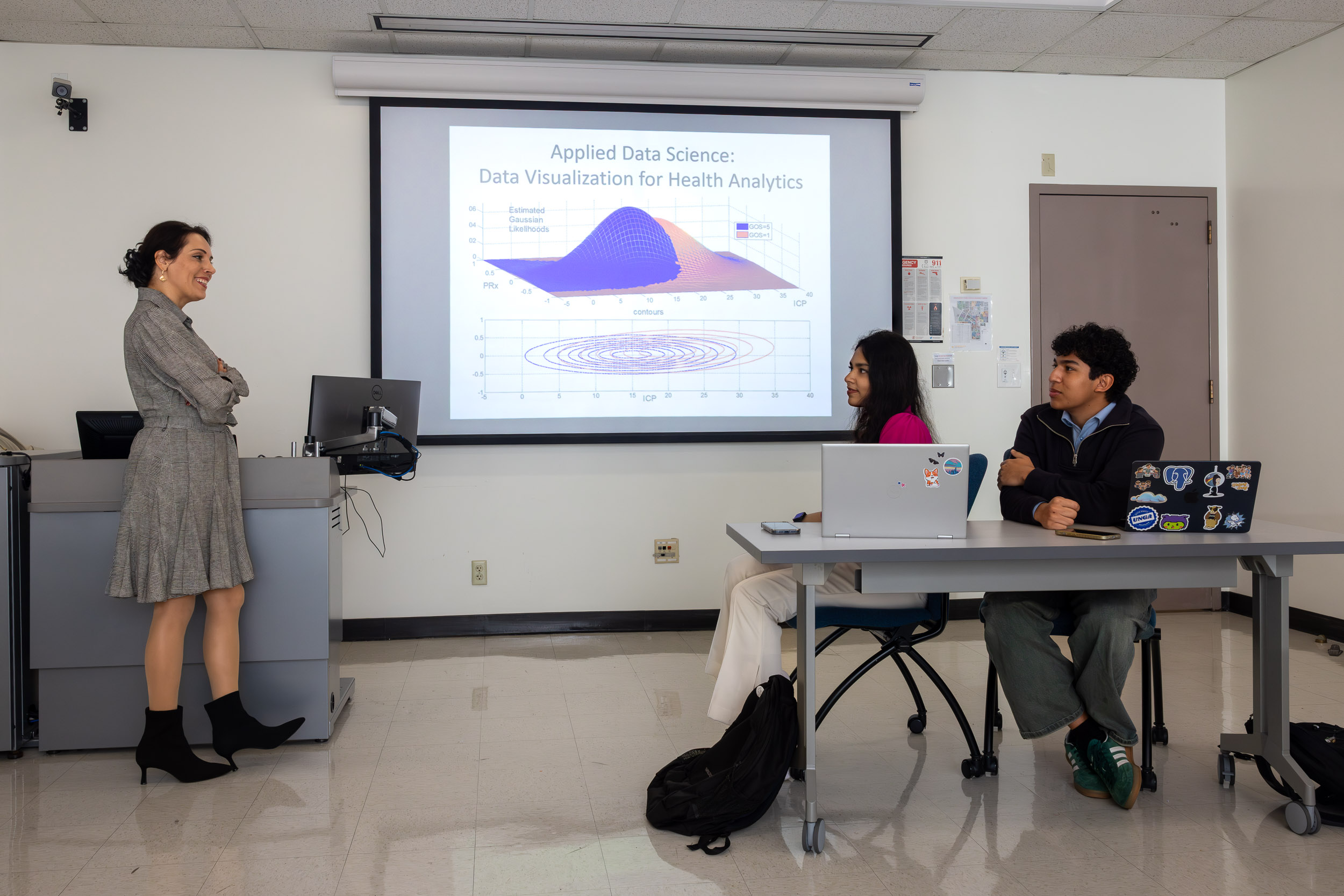 A person in a dress speaks with two seated individuals in a classroom