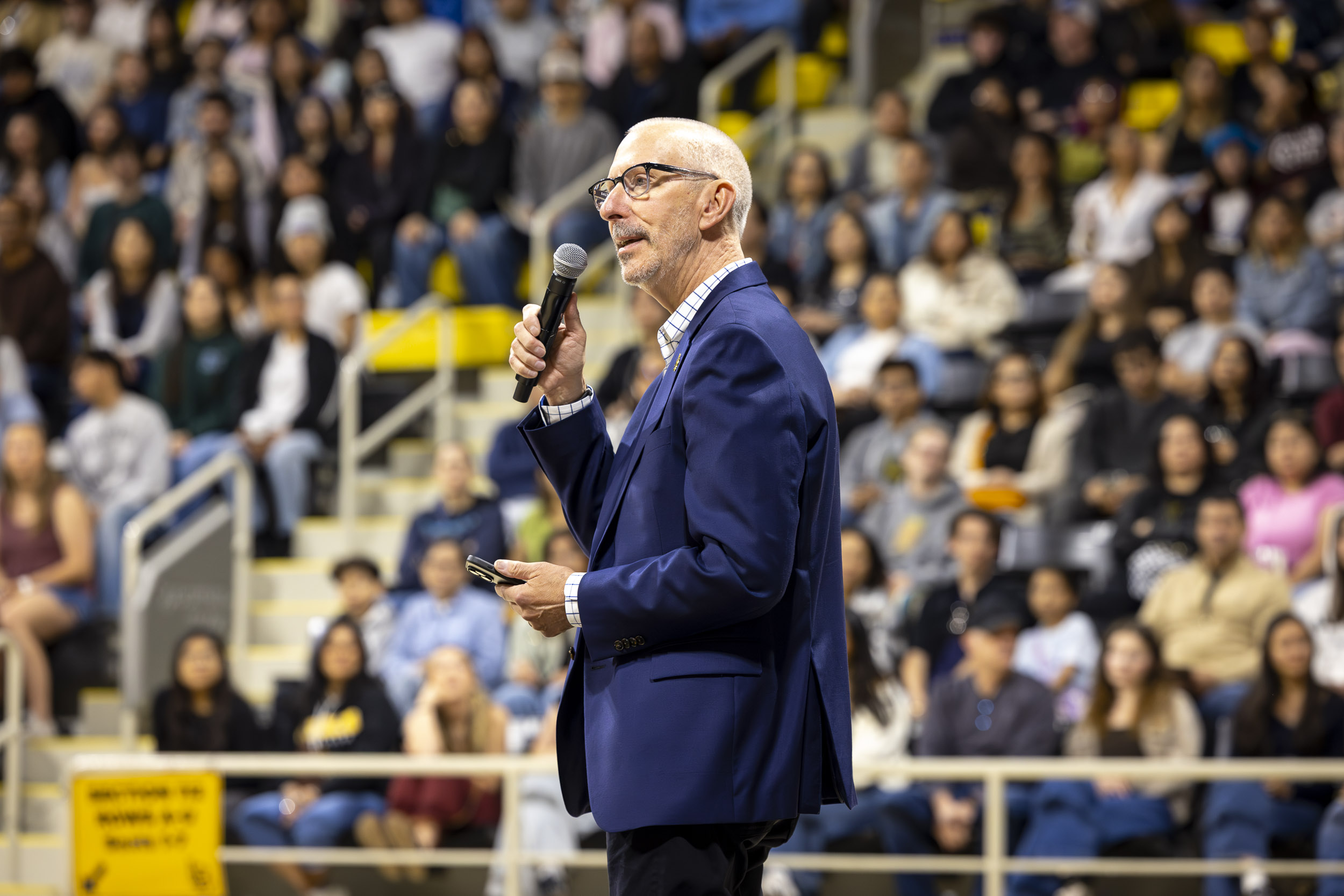Person in suit speaks into microphone while addressing seated audience in indoor arena