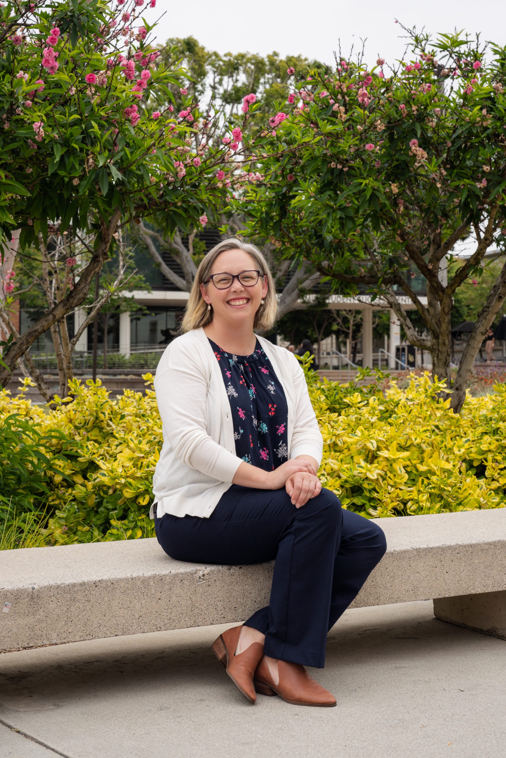 Dr Caitlin Fouratt seated on a bench. She has short brown hair, and is wearing blue pants, a blue floral blouse, white cardigan, and brown shoes.