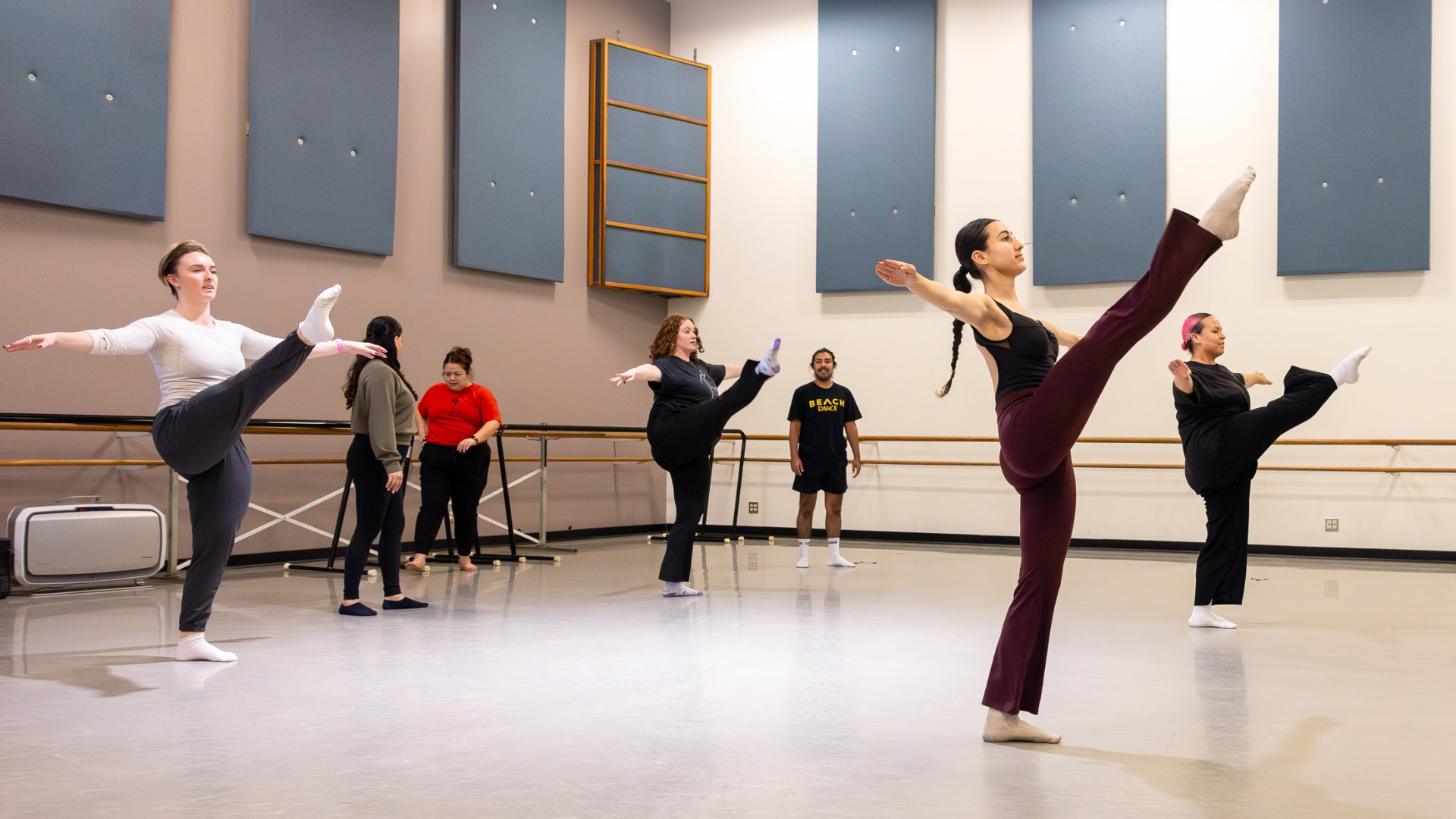 Dance students practice high kicks in a studio while an instructor watches from the back.