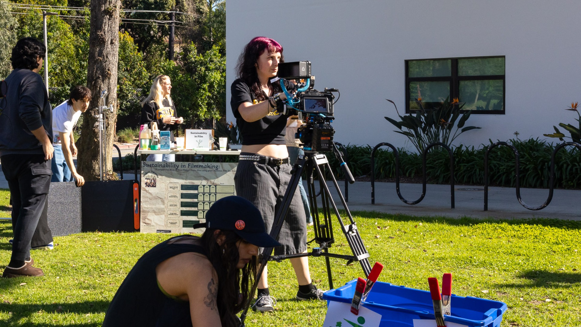 Student filmmakers work outdoors as one student operates a camera on a tripod.
