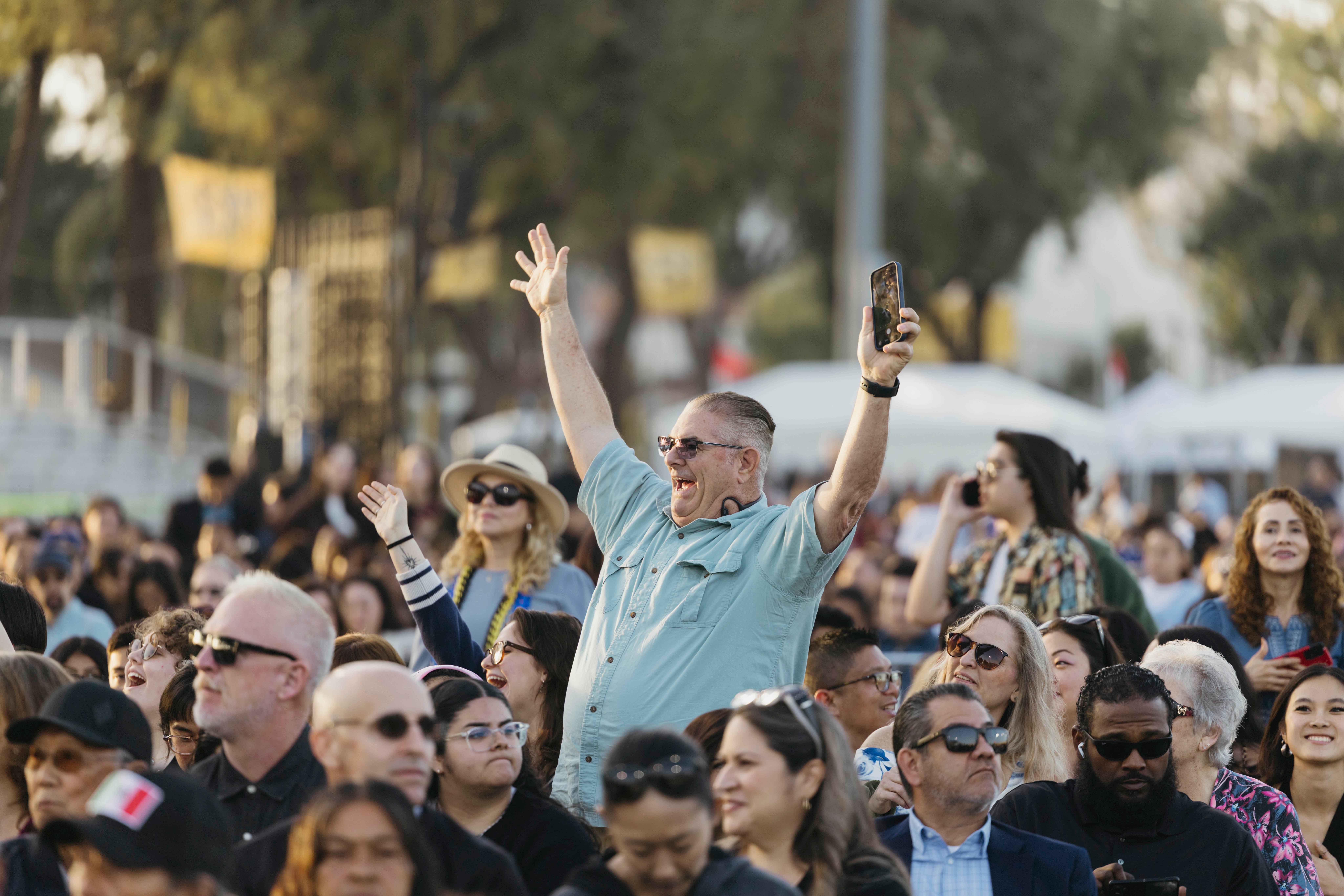 Person in crowd raises both arms cheering at outdoor graduation ceremony
