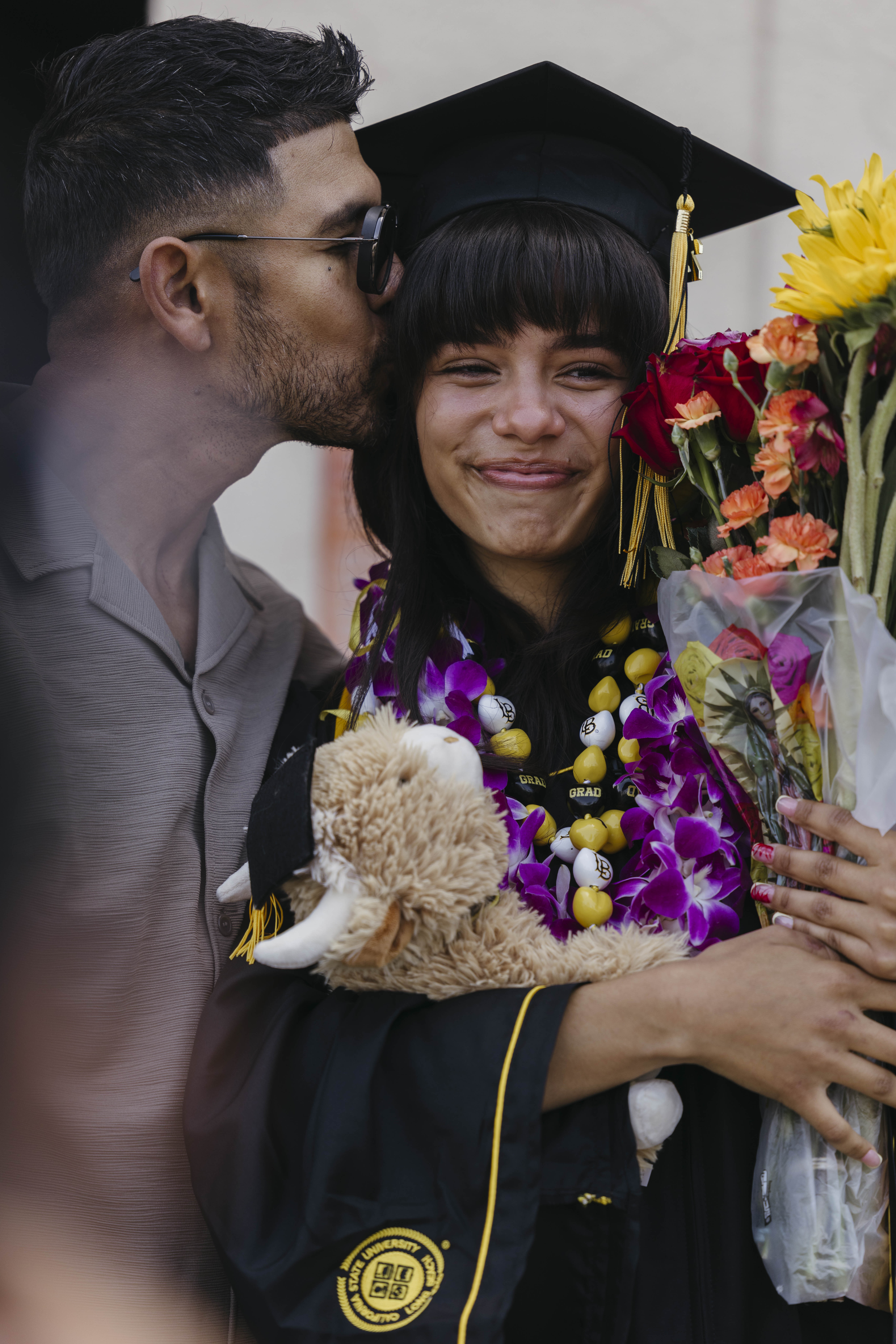 A person in cap and gown smiles while holding flowers and a stuffed animal