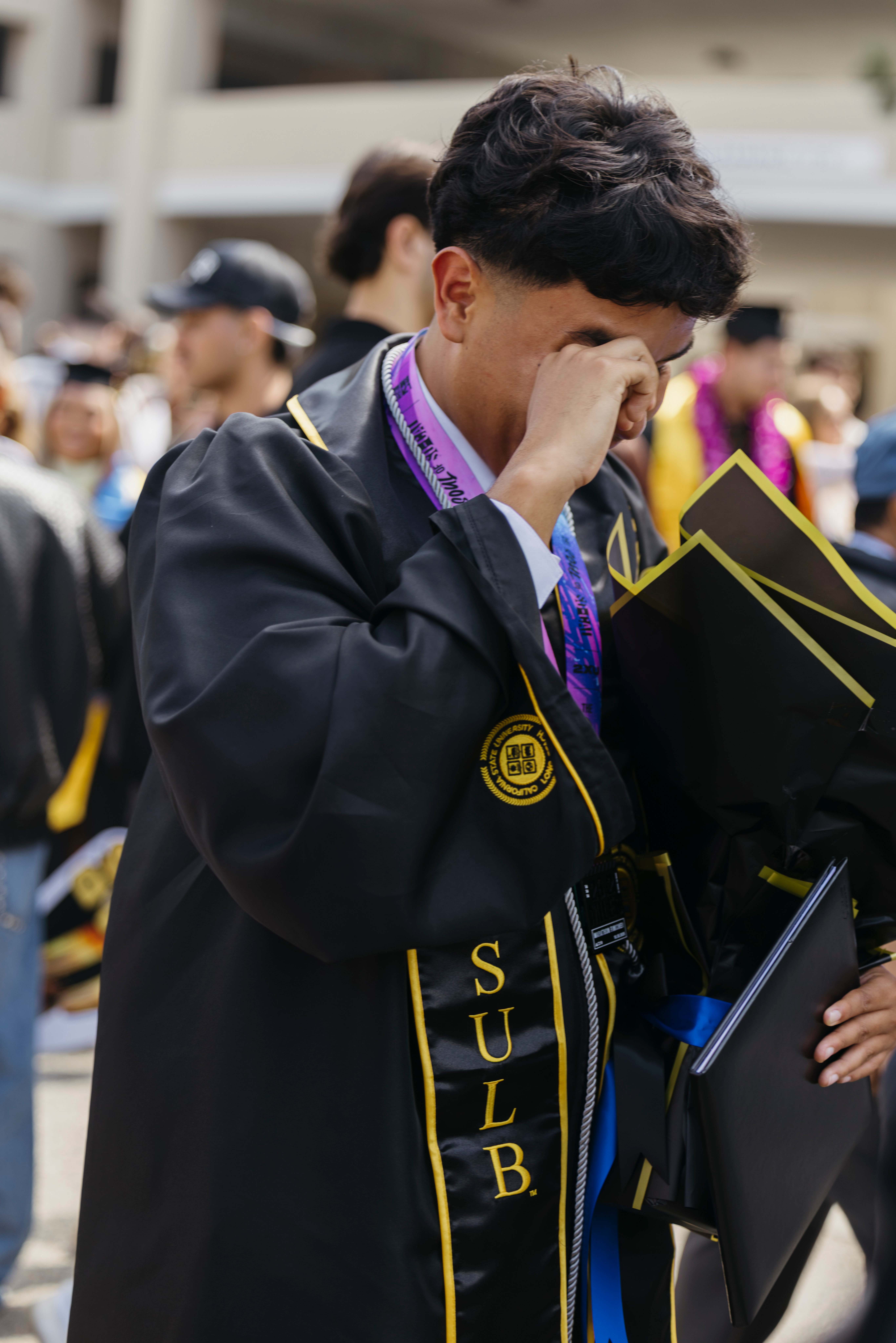Graduate in cap and gown wipes away tears while holding diploma