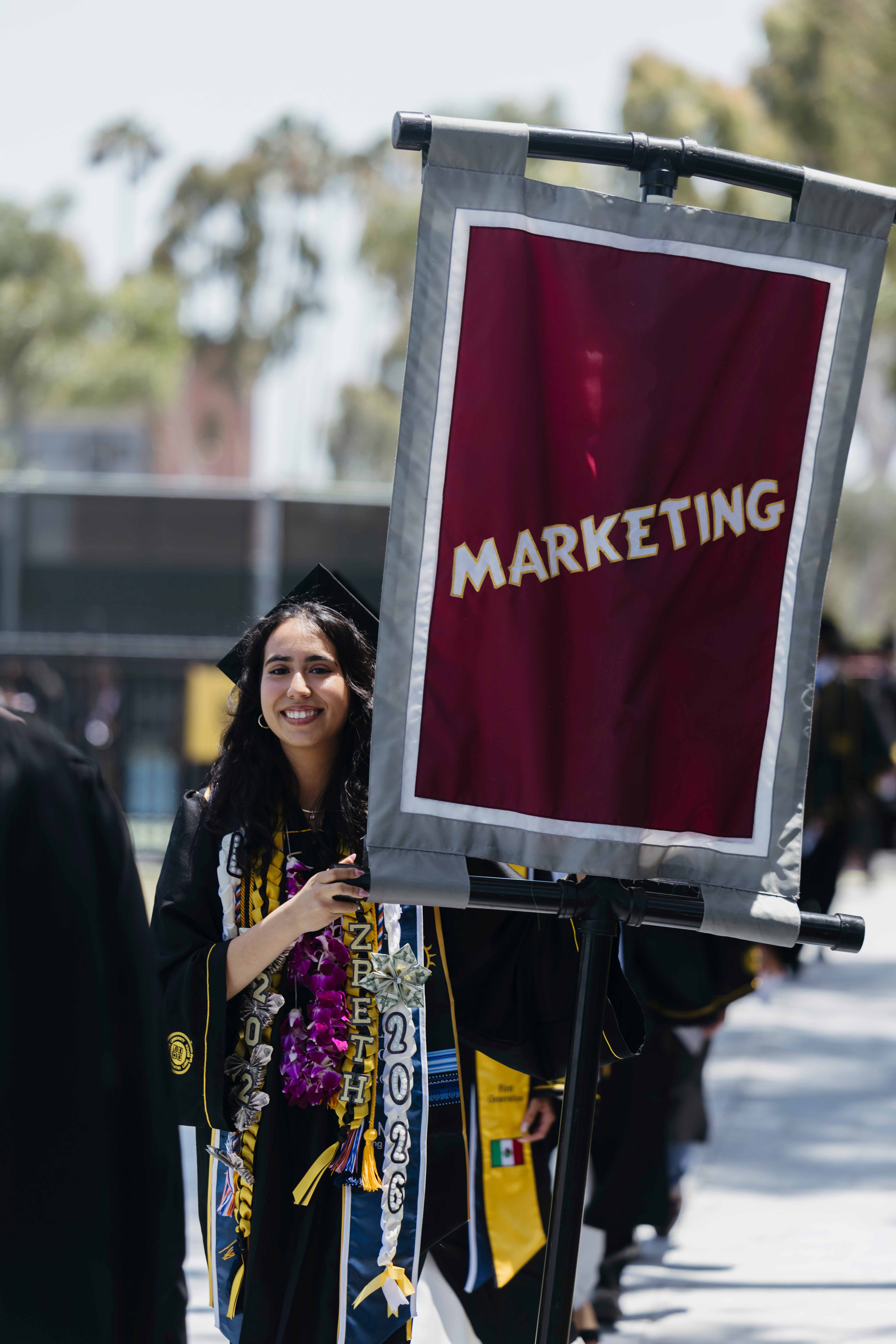 Graduate in cap and gown walks in procession next to Marketing banner