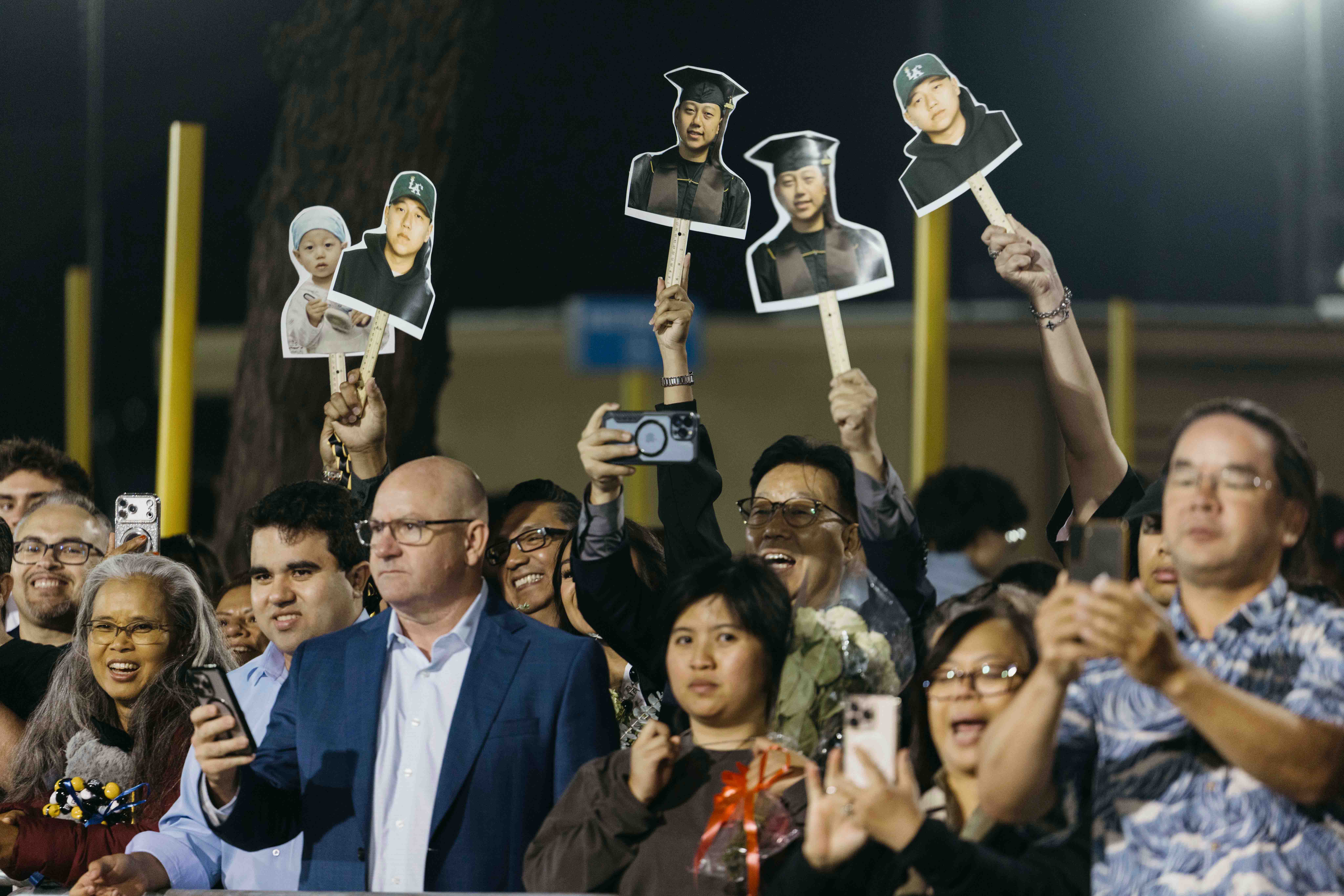 Crowd holds up large photo cutouts of graduate while cheering at night