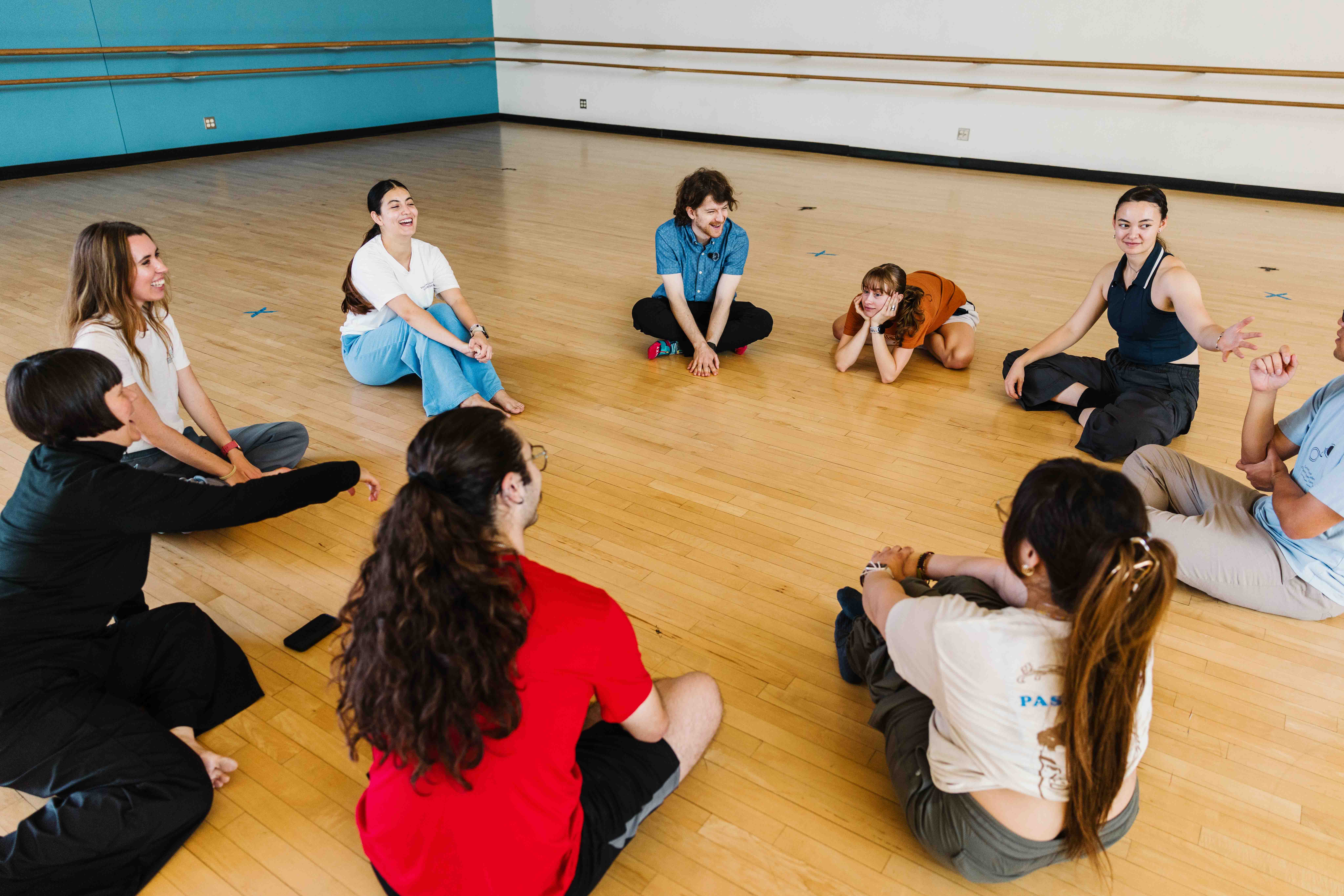 A group sits in a circle on a dance studio floor, laughing and talking