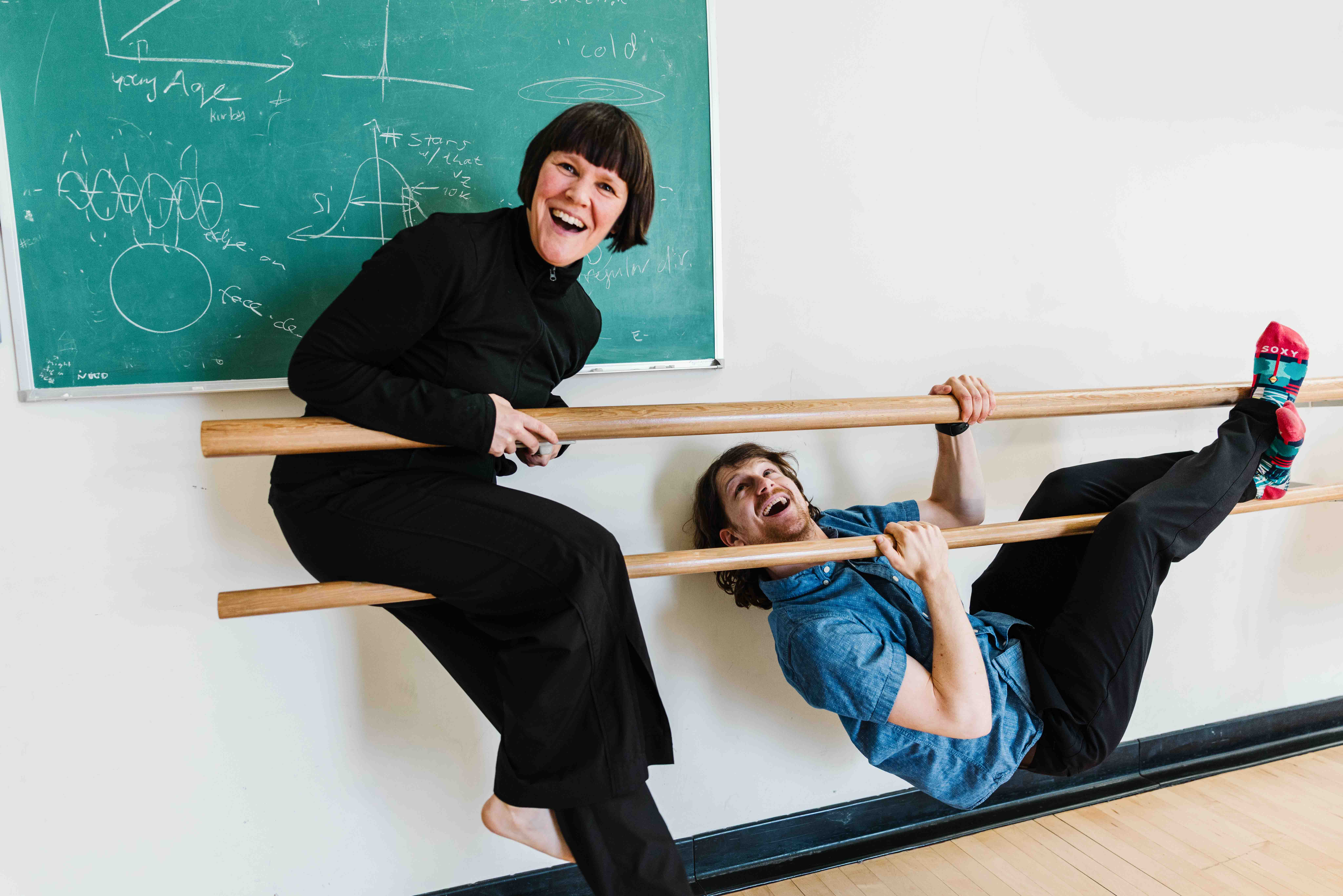 Two people laugh while perched on a ballet barre in a dance studio