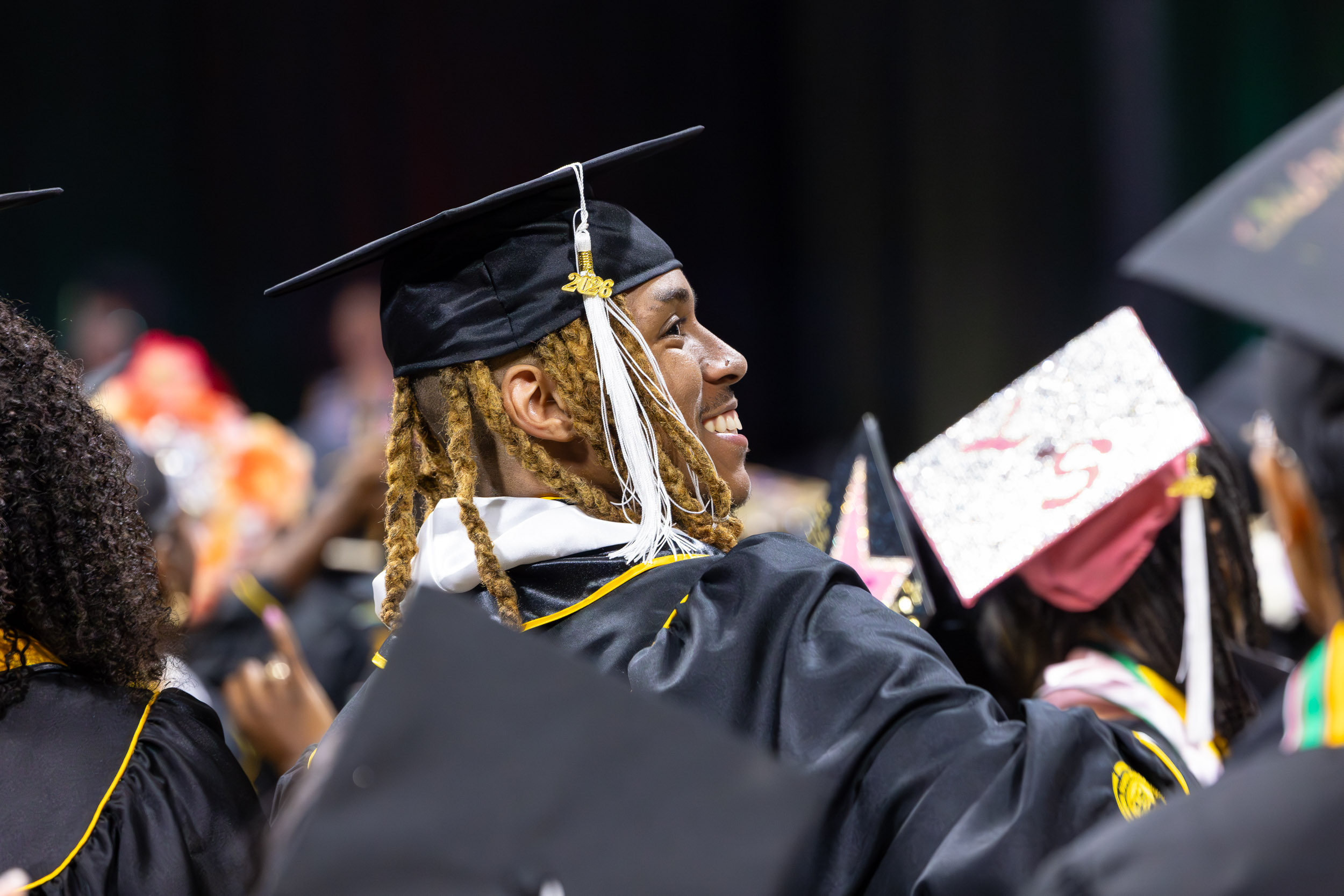 A person in cap and gown smiles during an indoor graduation ceremony