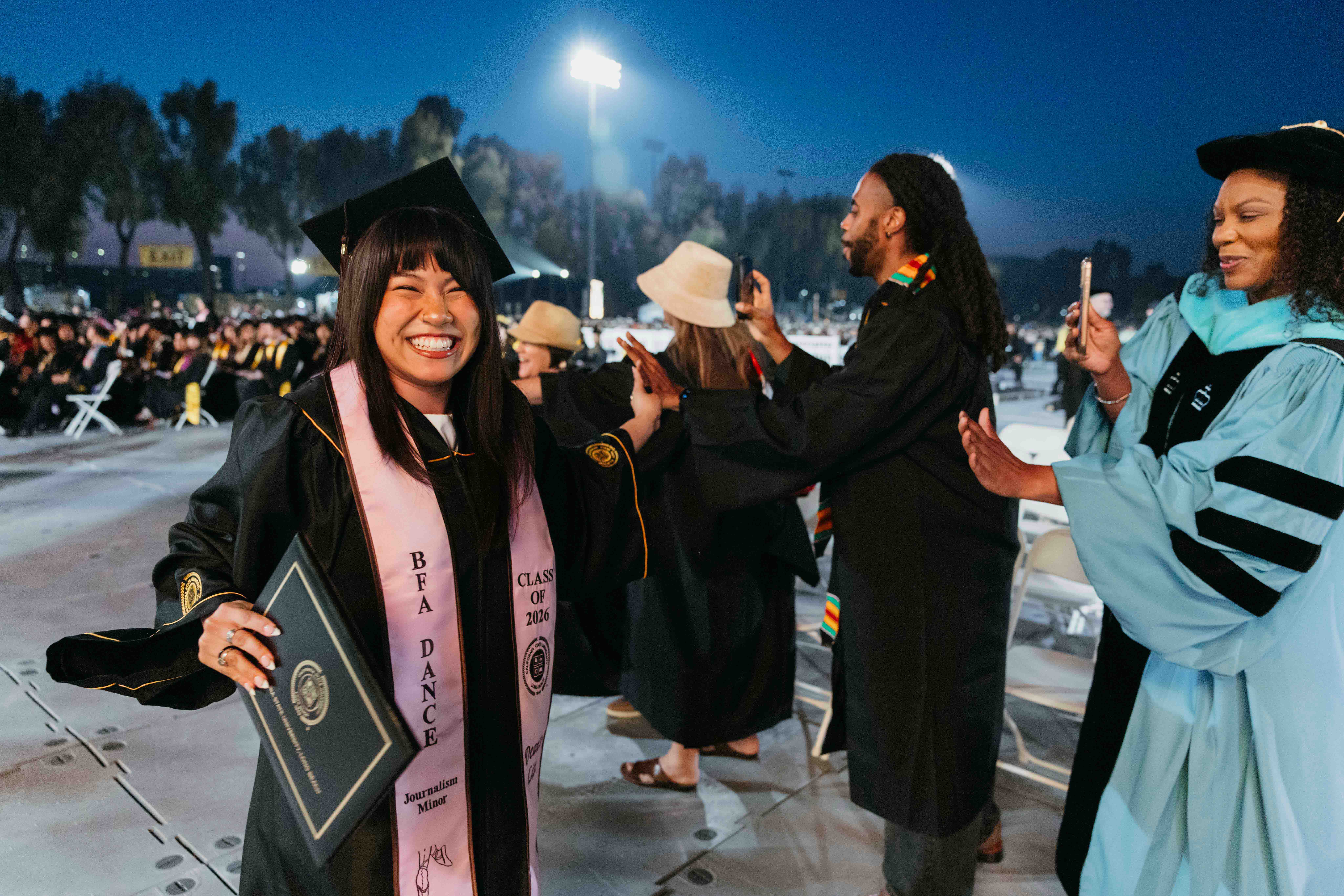 Person in cap and gown smiles holding diploma at evening ceremony