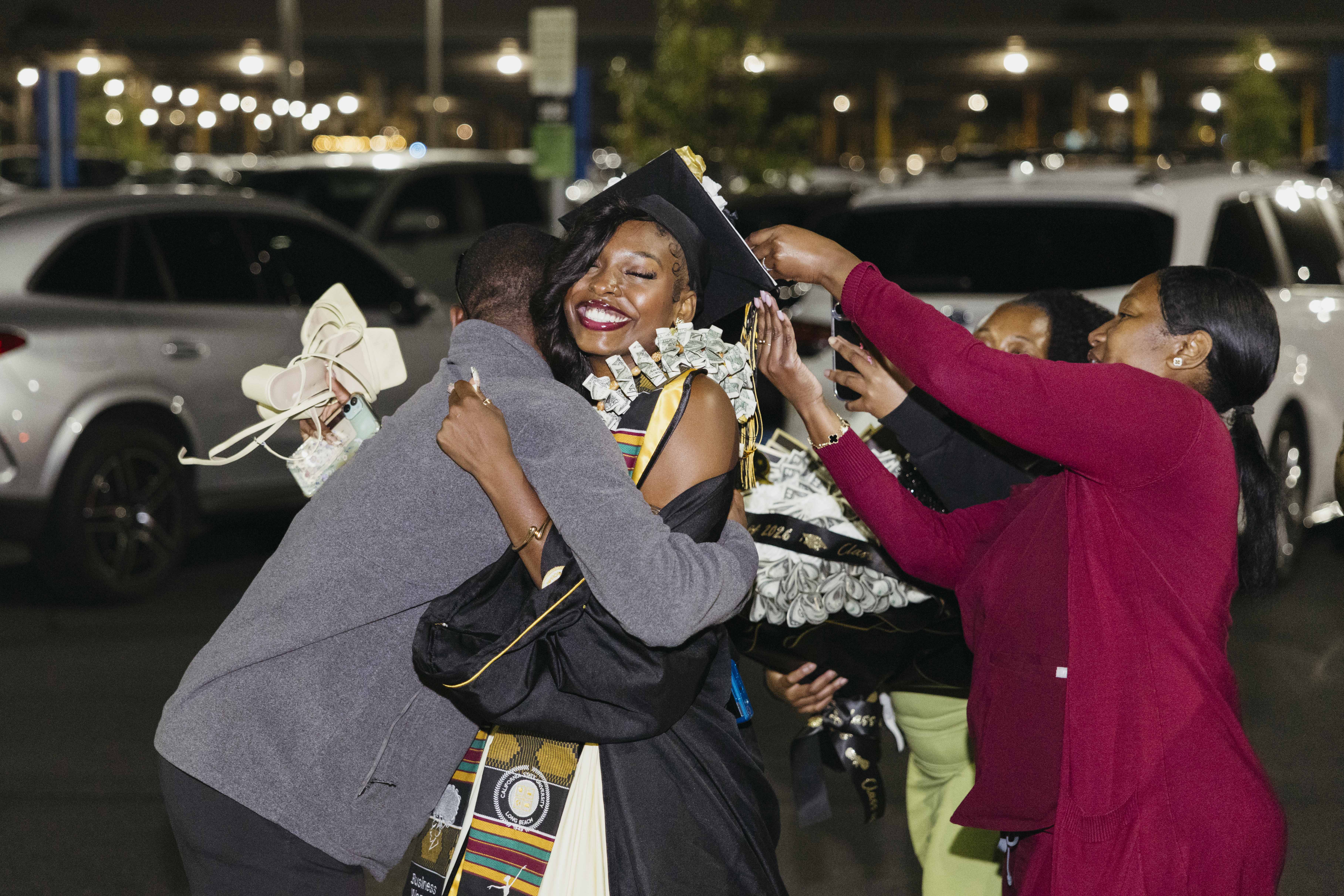 Graduate in cap and gown hugs family members in parking lot at night