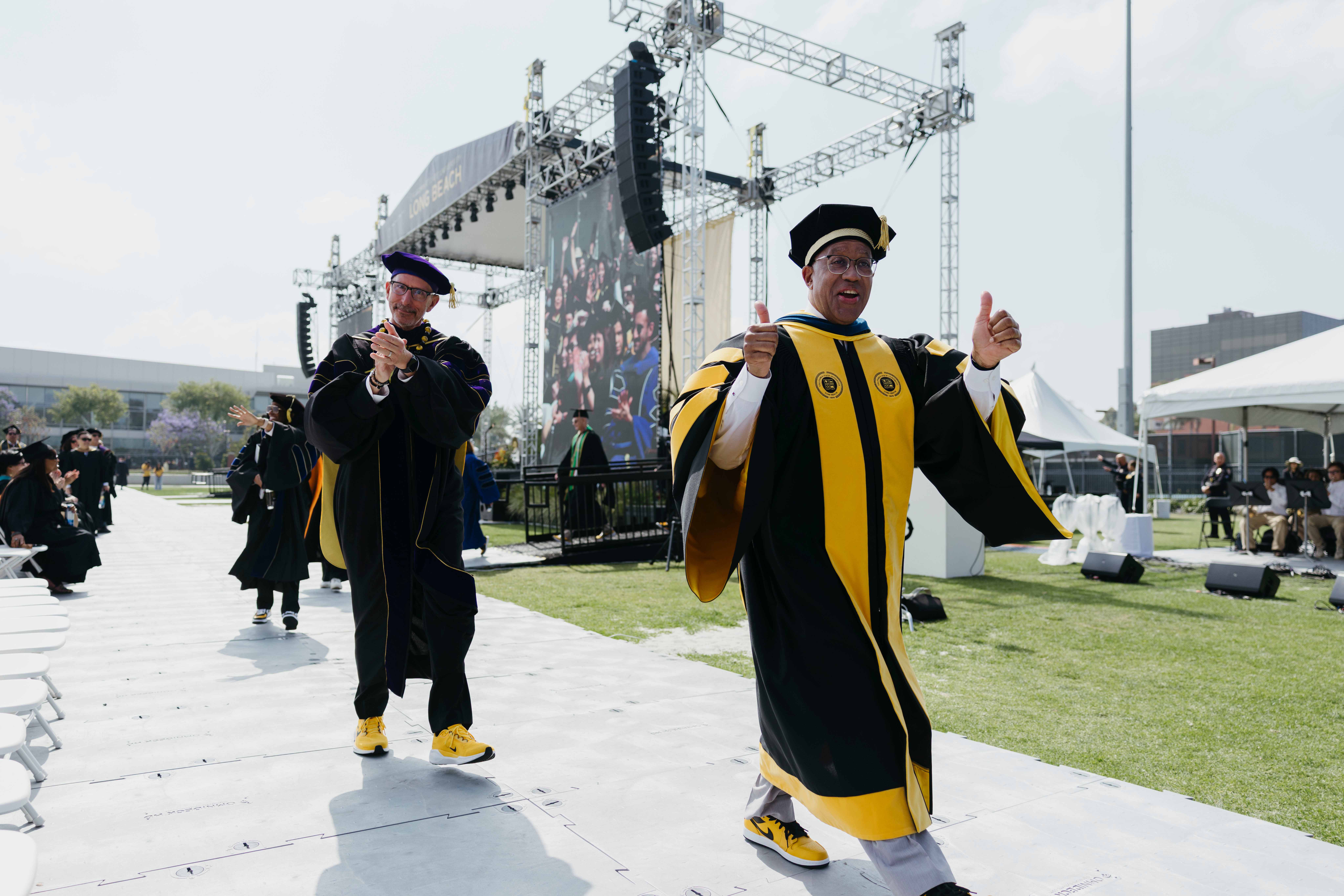 Two people in academic regalia give thumbs up while walking down aisle at outdoor ceremony