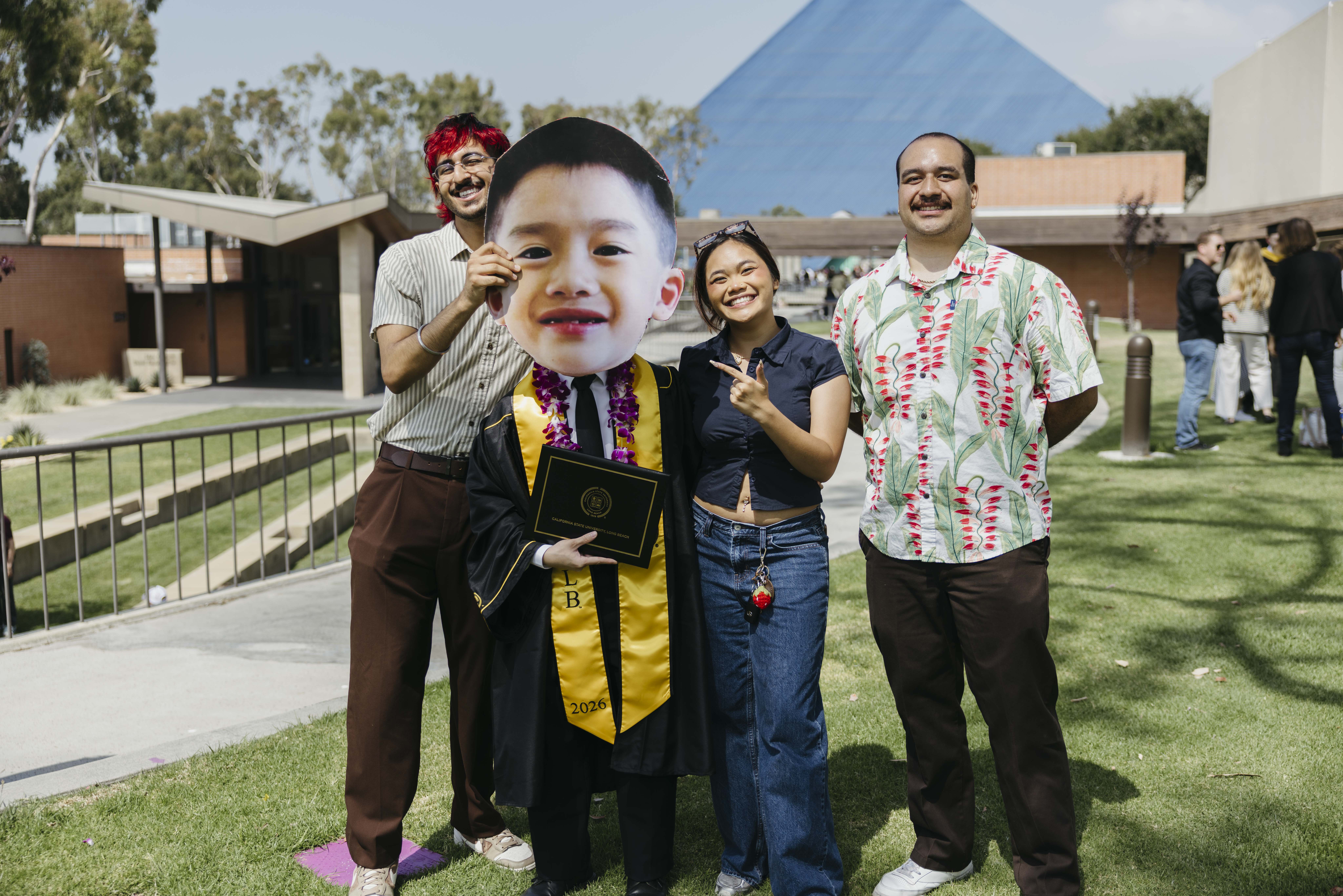 Graduate poses with group holding oversized face cutout on campus lawn