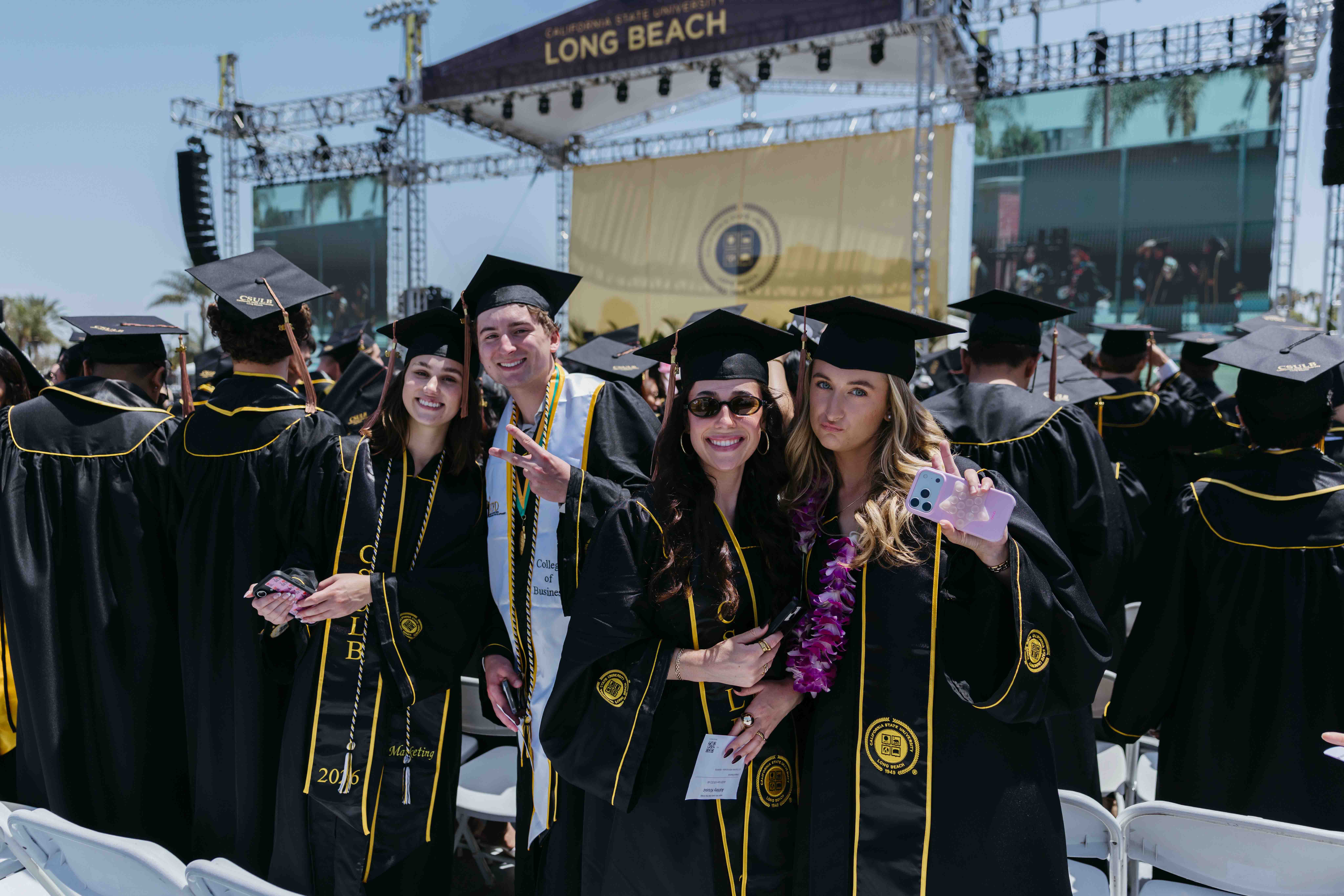Four graduates pose together in front of CSULB commencement stage