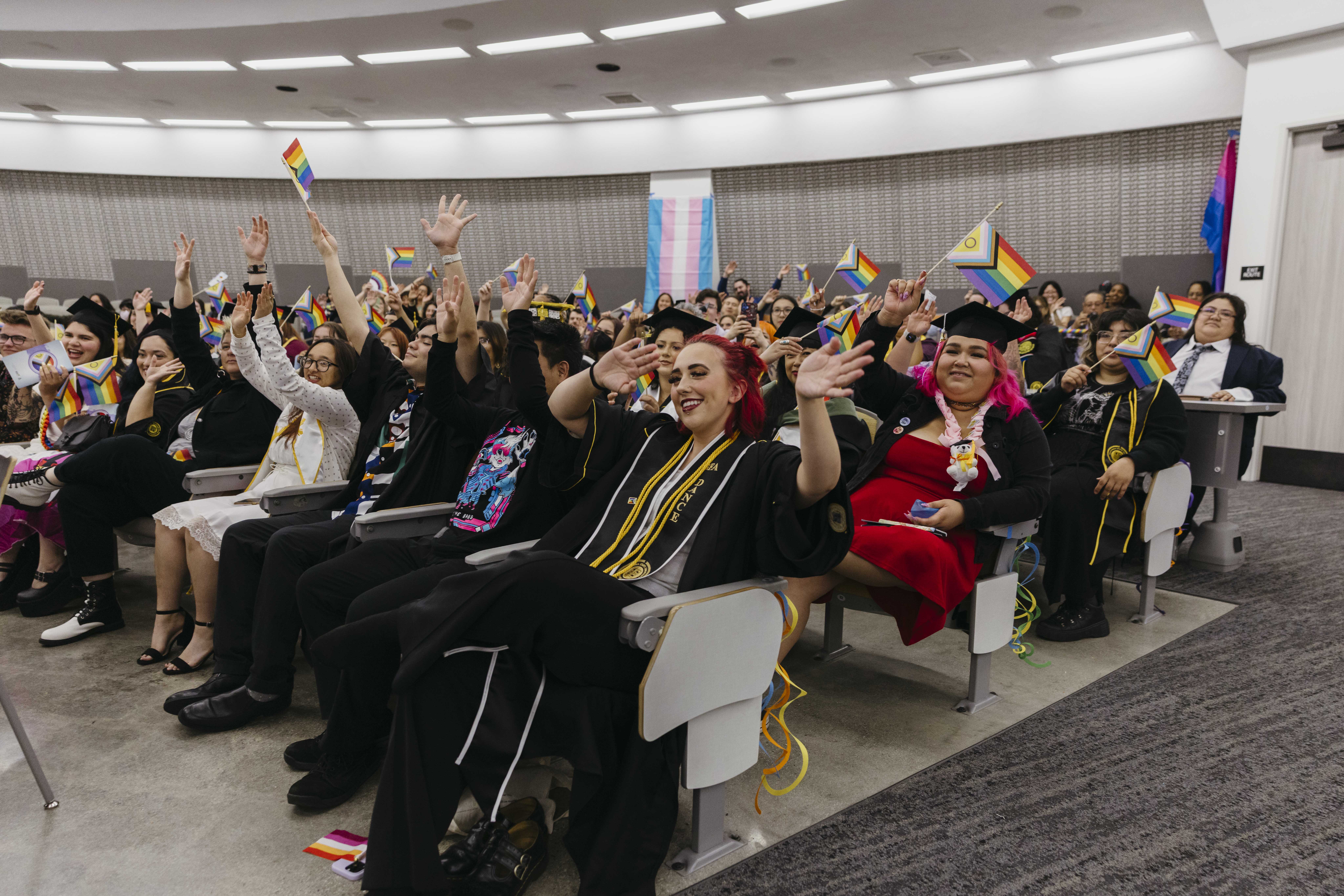 Crowd of graduates raise hands and wave pride flags at indoor ceremony