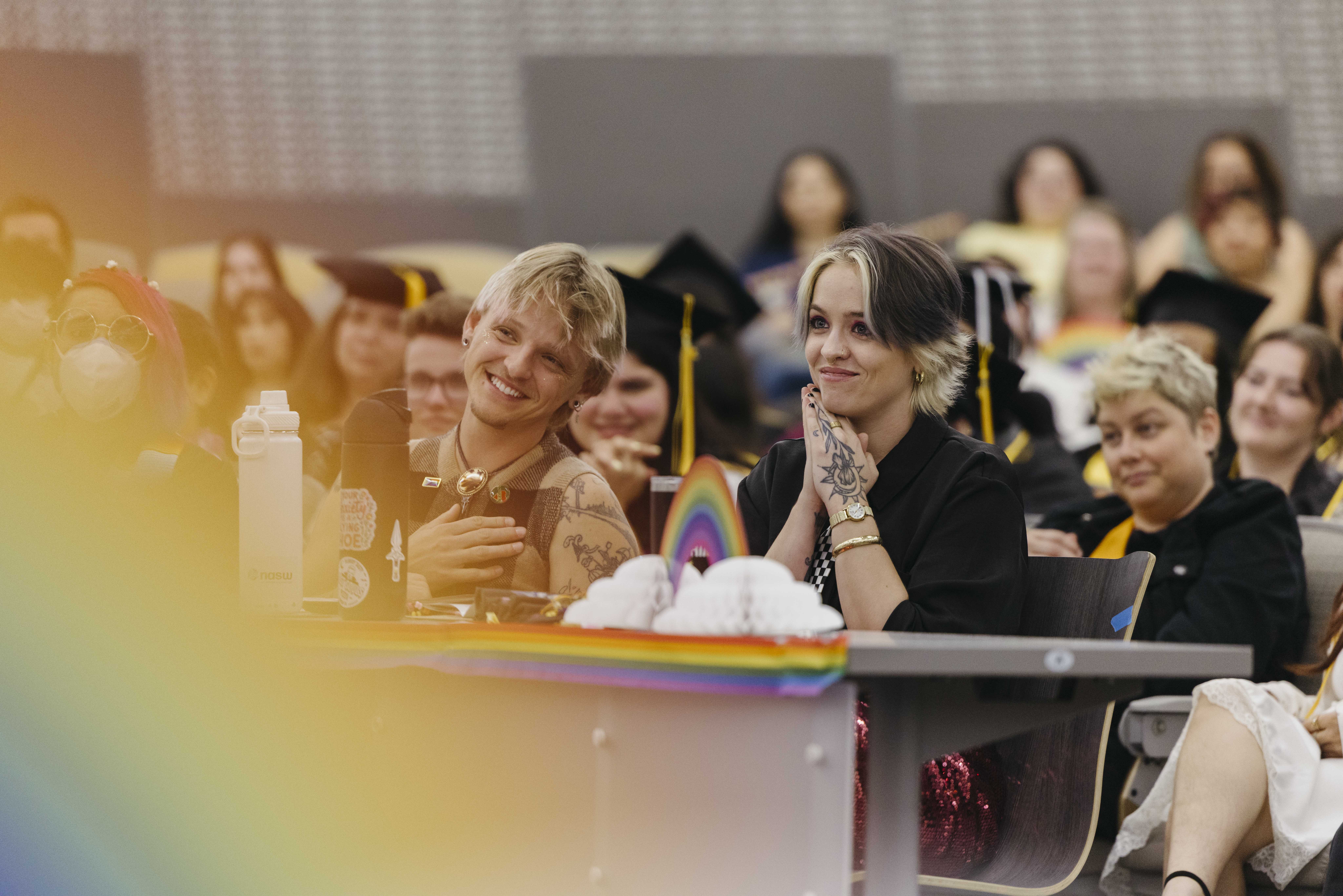  Two people in graduation attire smile during indoor ceremony with pride decorations