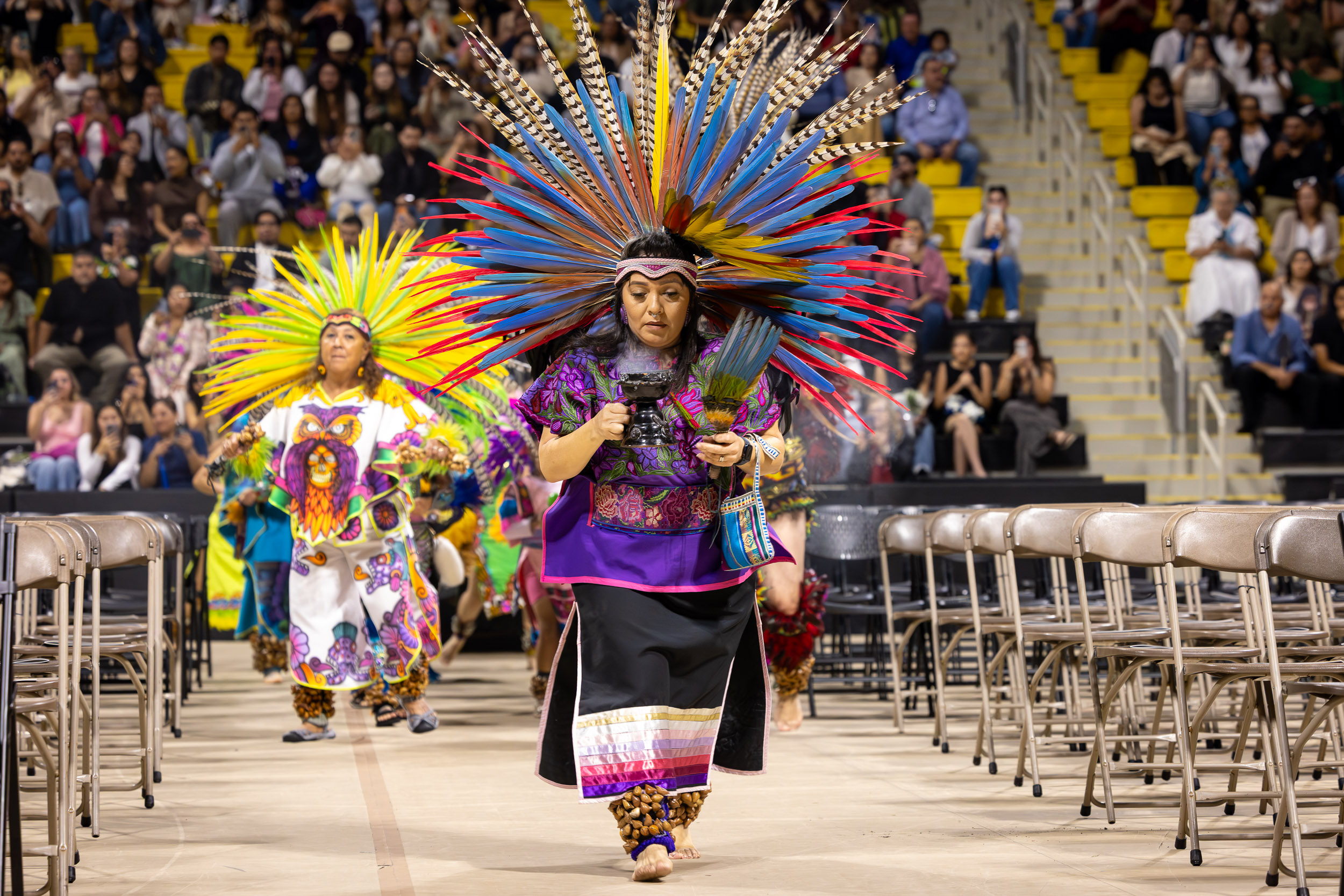 Performers in colorful traditional regalia and feathered headdresses process down aisle at indoor ceremony