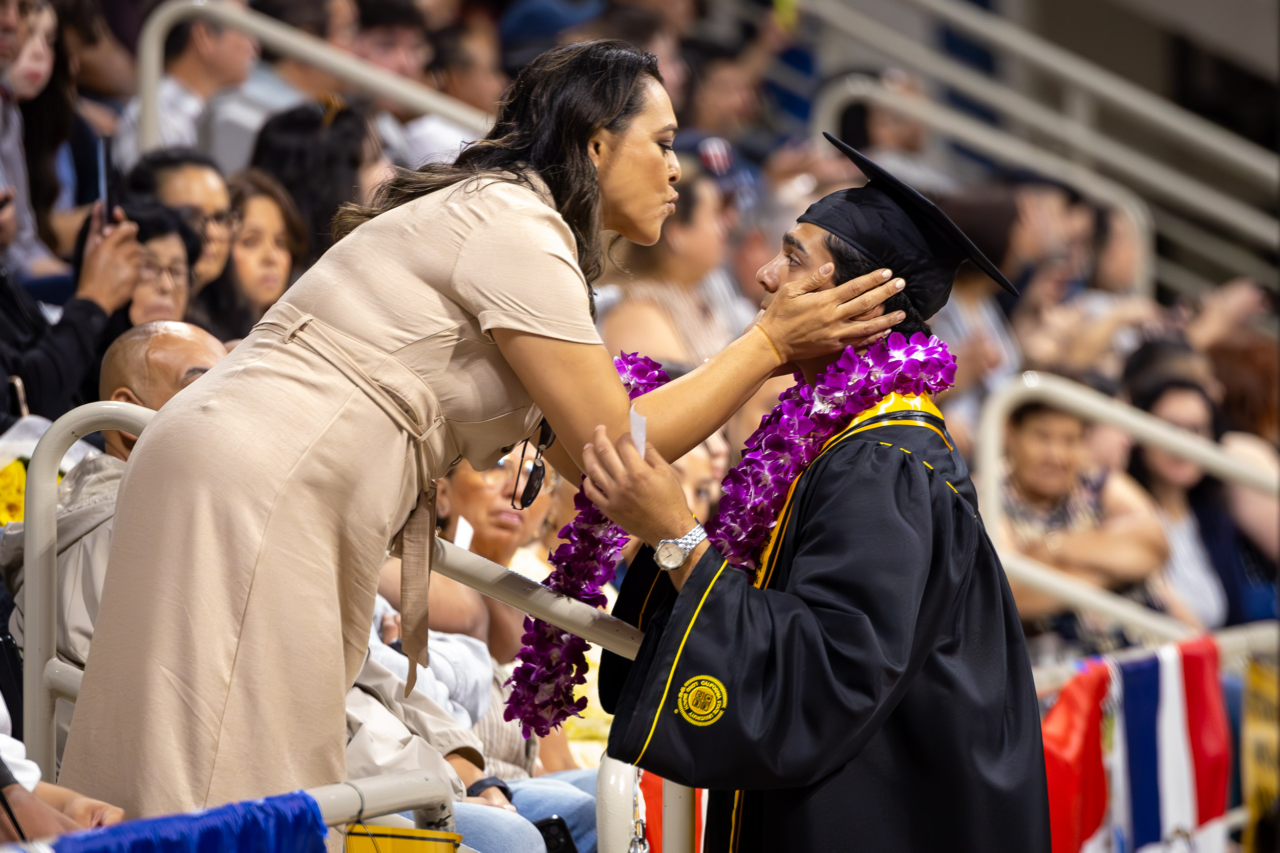 A person leans over a railing to kiss a graduate on the cheek at an indoor ceremony