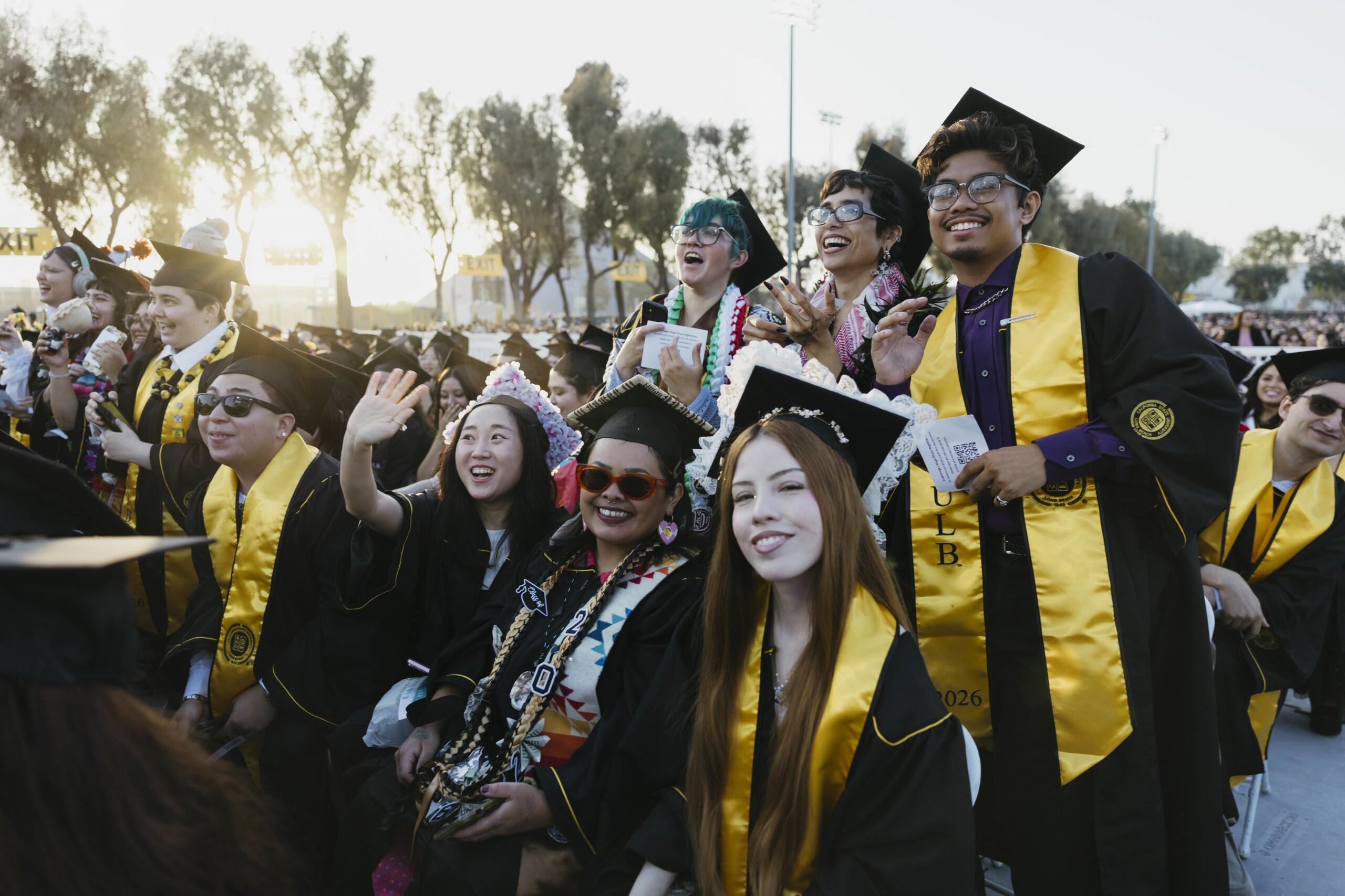 Group of graduates laugh and wave in caps and gowns at outdoor ceremony
