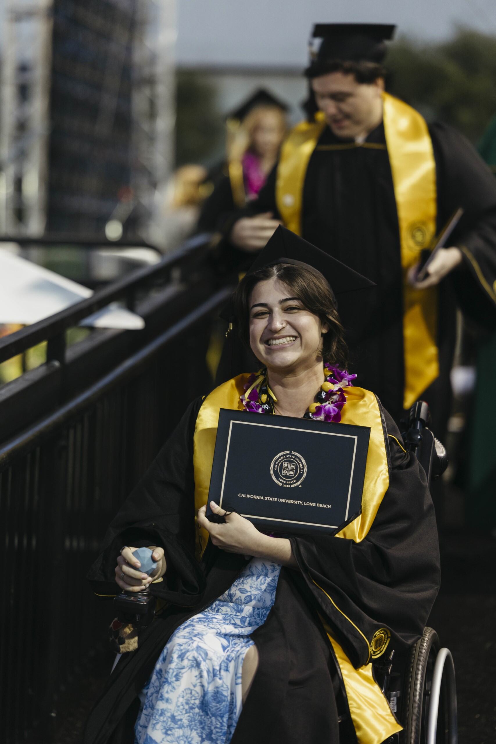 A person in graduation regalia holds a diploma while using a wheelchair on a ramp