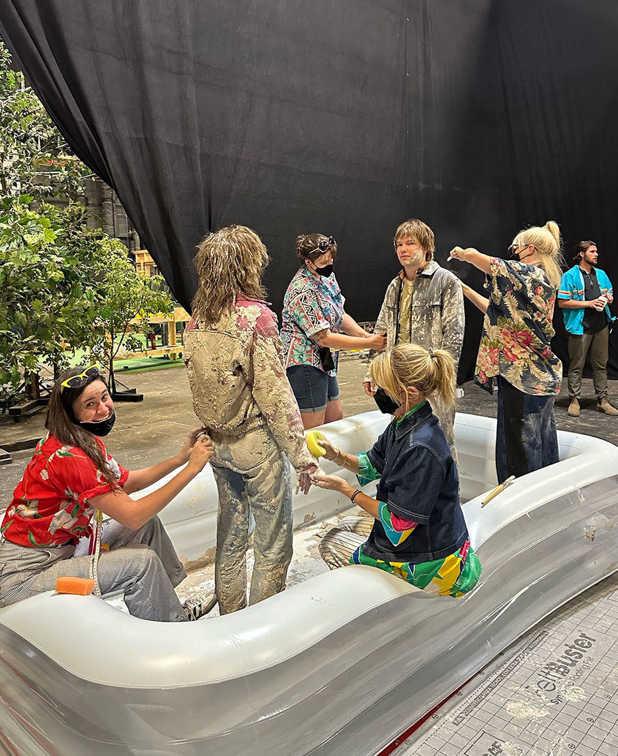 Actors covered in mud stand in an inflatable pool with crew members