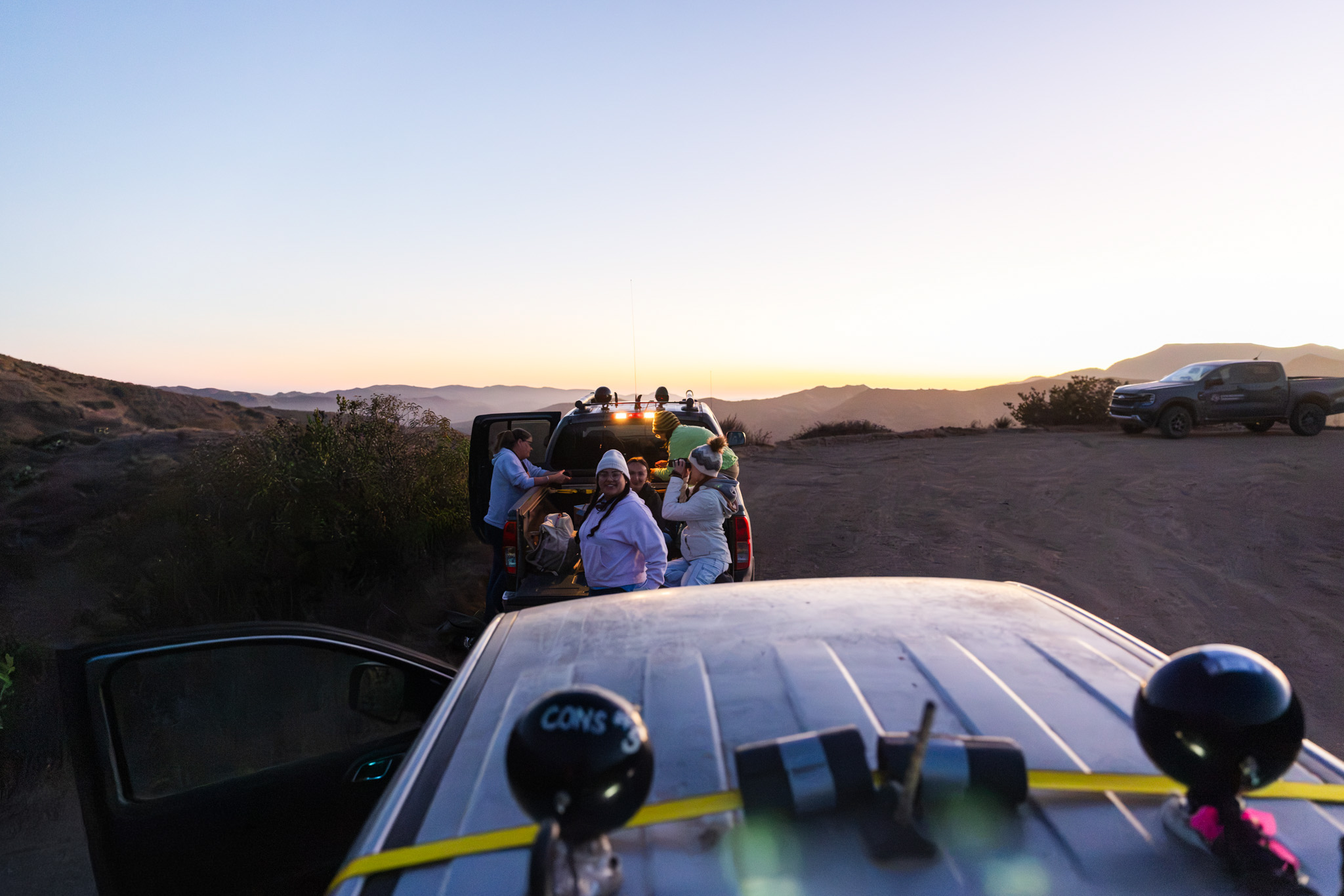 A group gathers in and around truck beds on a dirt road at sunset with spotlights mounted overhead