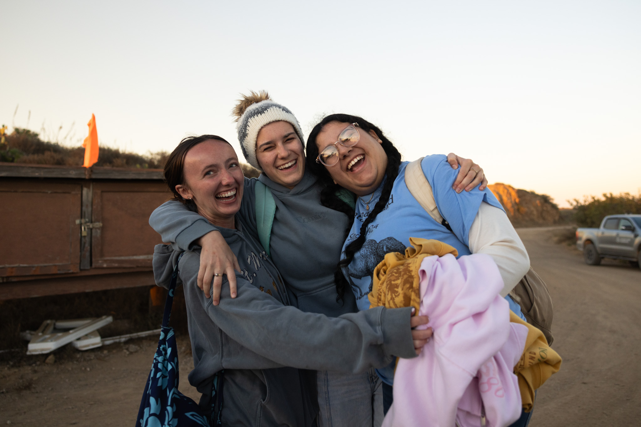 Three people embrace and laugh together outdoors at sunset
