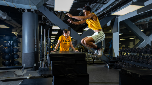 Two athletes practicing in a gym, with one athlete jumping over a stack of padded boxes while another watches.