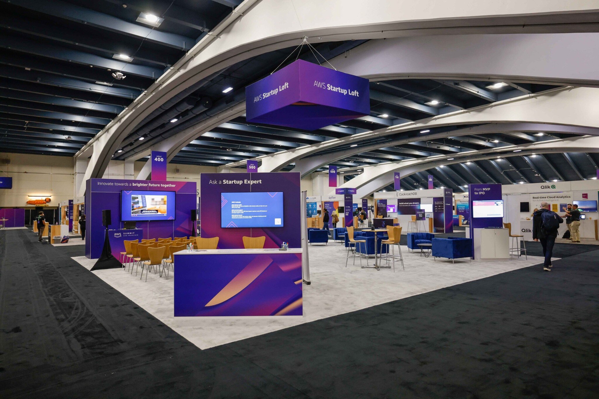 Purple AWS Startup Loft exhibit with presentation screens, seating, and consultation areas at a conference. Created by Taylor Gibbs