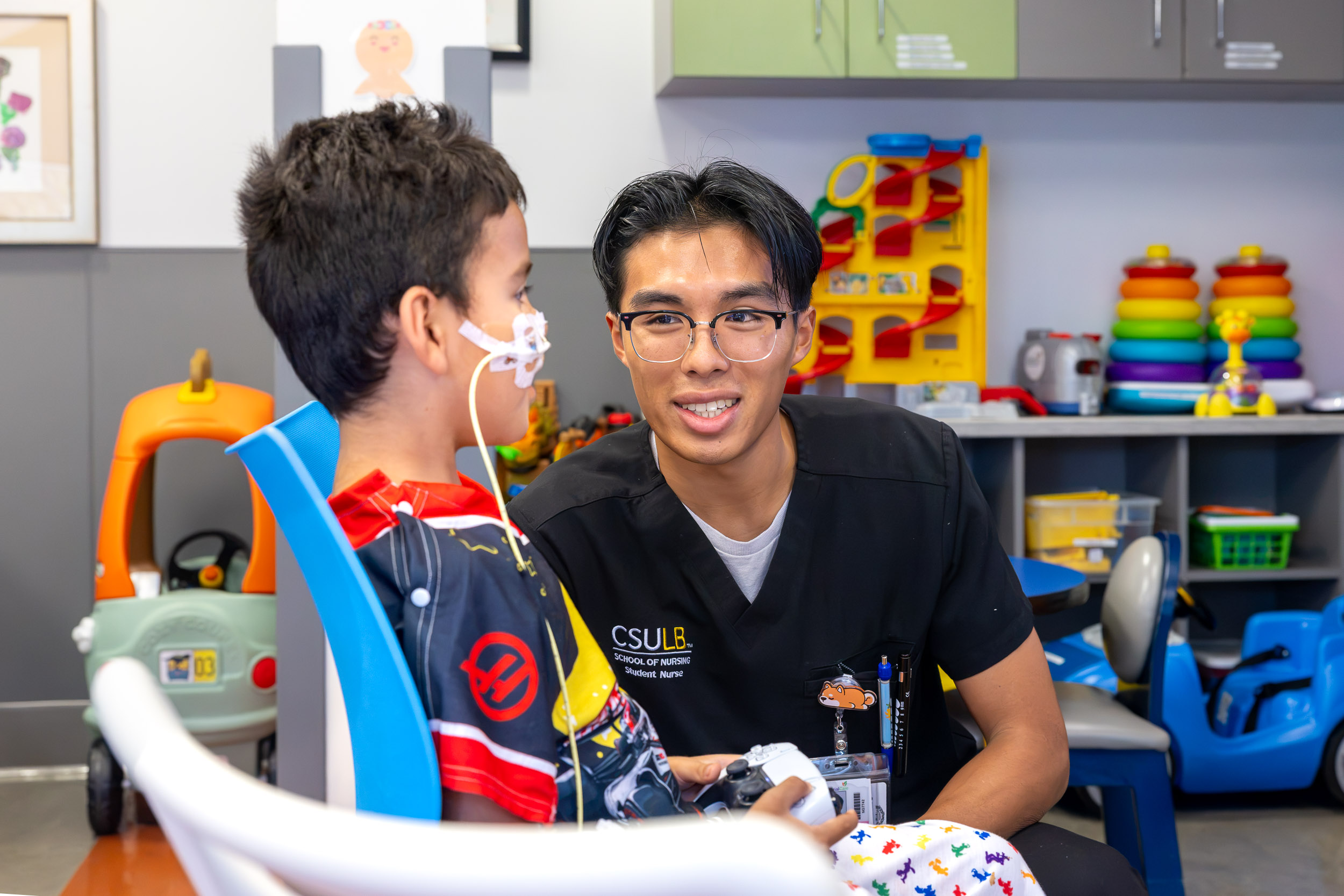 A person in scrubs smiles at a young patient holding a game controller.
