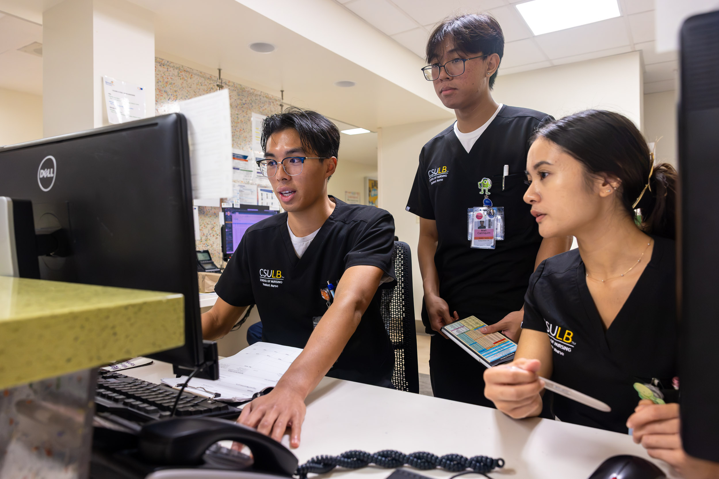 Three people in scrubs look at a computer at a hospital nurses’ station.