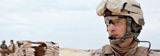 Three military men stand in front of a sandbag shelter while another man in military gear gazes over the desert