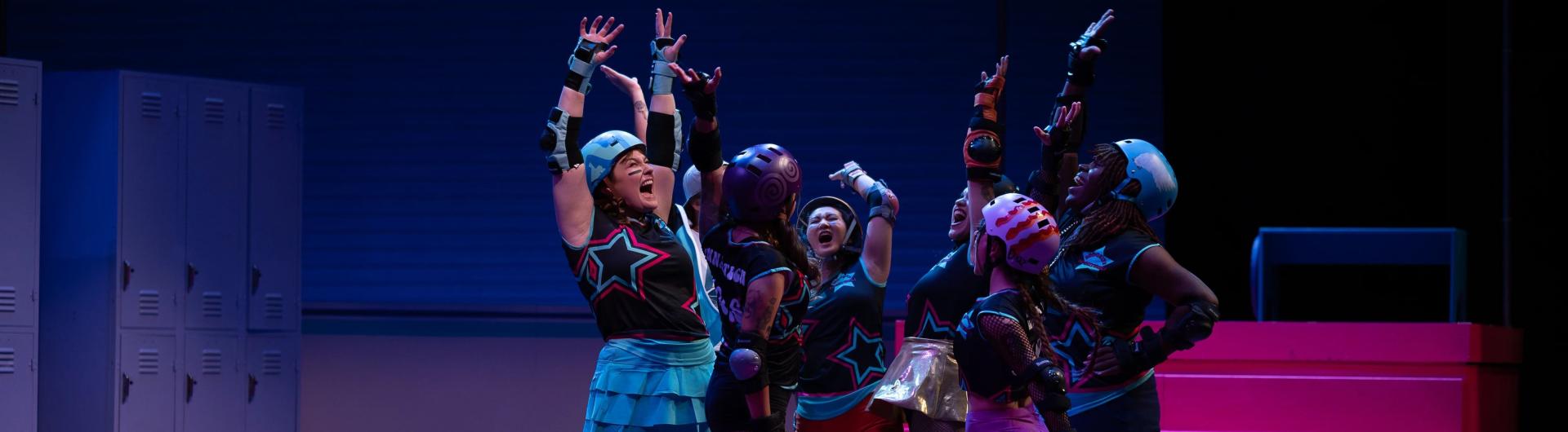 Theatre students performing Roller Derby Play; a group of actors wearing roller derby gear have their hands up in the air.