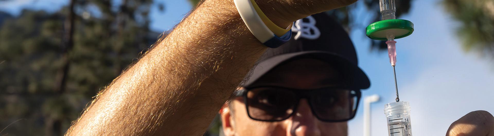 A person holds a syringe over a clear tube, releasing drops of liquid during outdoor sample collection.
