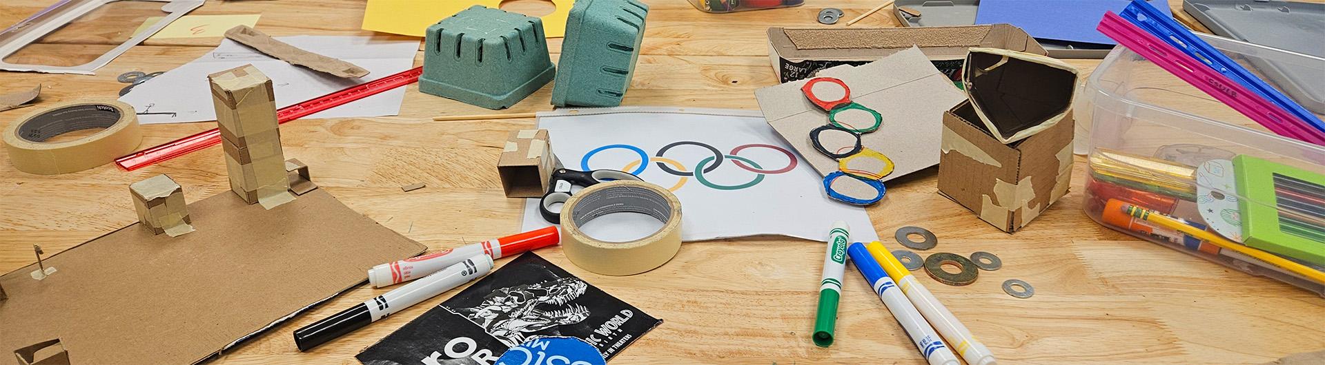 markers, paper, cardboard, tape, and other craft objects strewn across a table