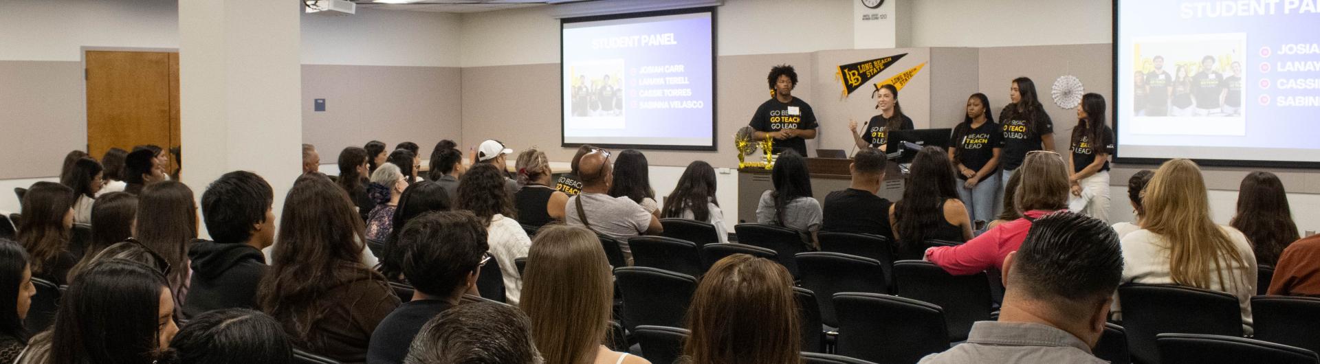 A crowd listens to a group of students speak as a panel in the front of a large room