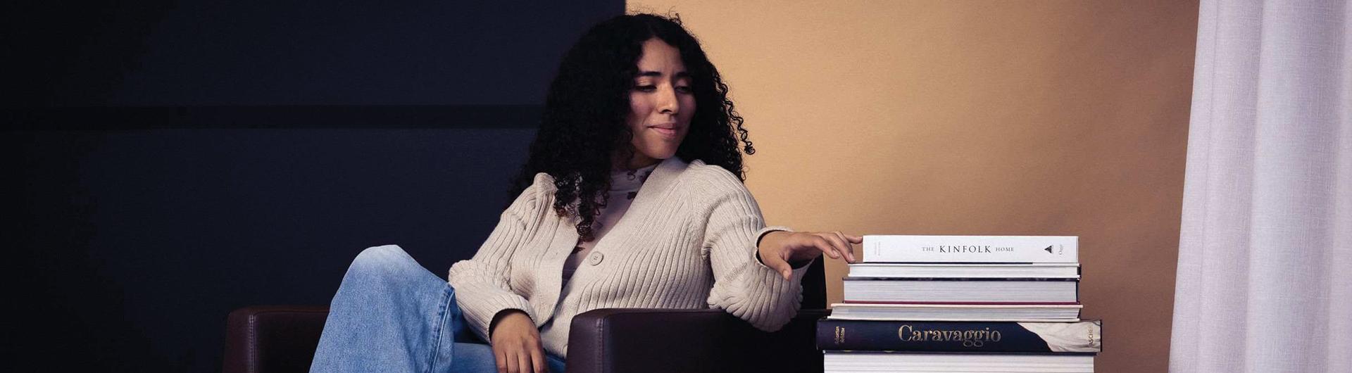 A person sits on a chair and looks at a stack of books next to her