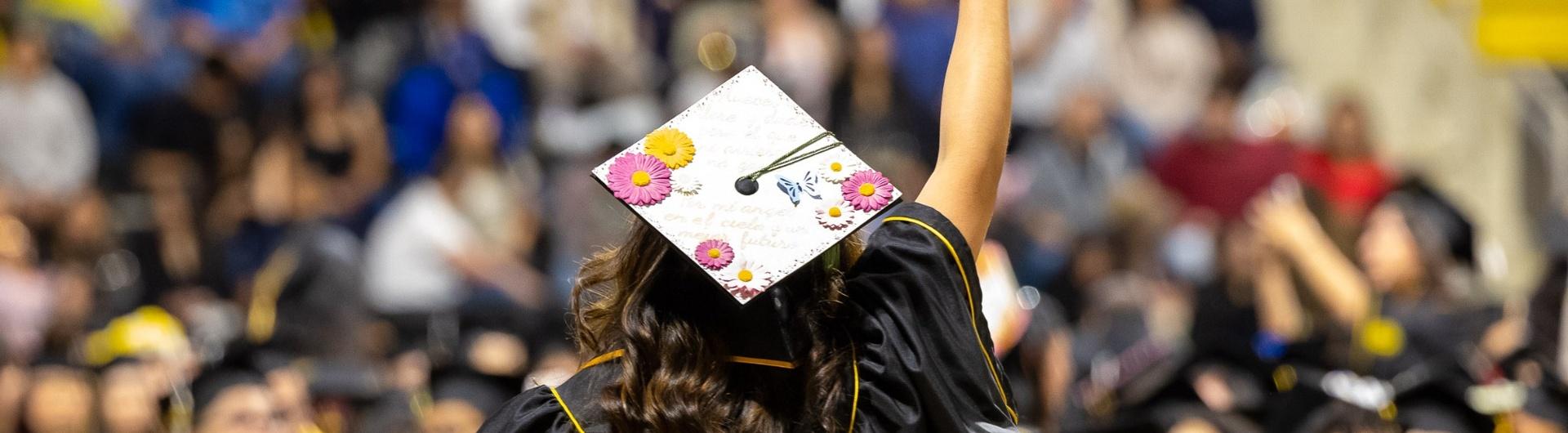 An image of a person in a cap and gown from behind with their hand rasied towards a crowd of people.