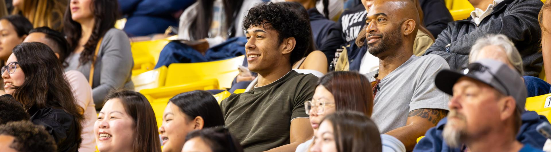 A large, diverse audience sits in bright yellow bleachers inside an indoor arena. Several spectators are smiling and watching an event, while others are talking or looking toward the floor. In the center, a young man in a dark green shirt smiles beside a man in a gray T-shirt. The foreground and background are softly blurred, emphasizing the central spectators.