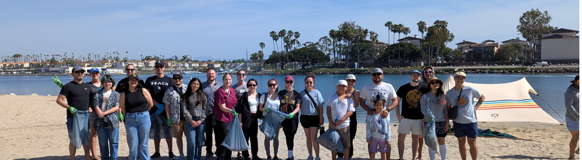 Beach cleanup volunteers pose with trash bags on a sunny shoreline with palm trees and water in the background