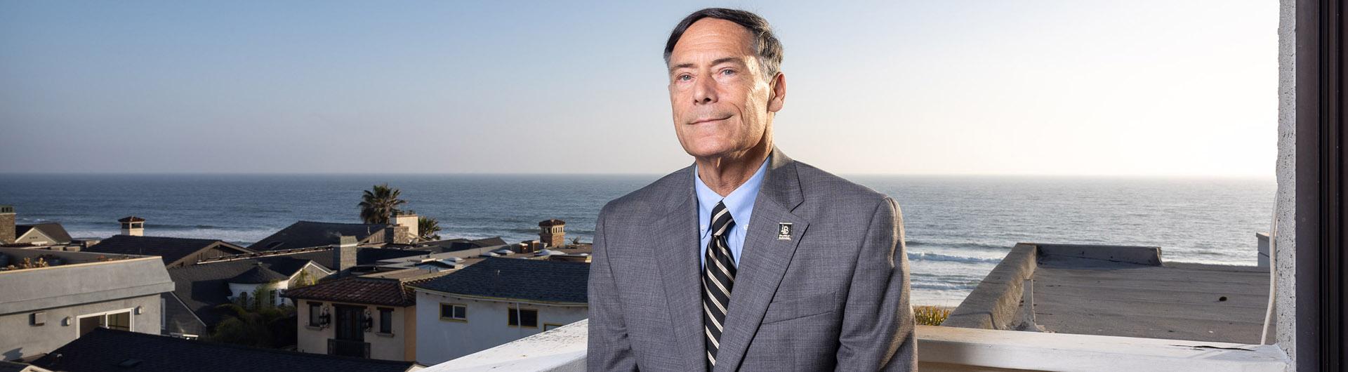 Wayne Powell faces the camera with a group of residences behind him, and the Pacific Ocean and the beach behind that area.