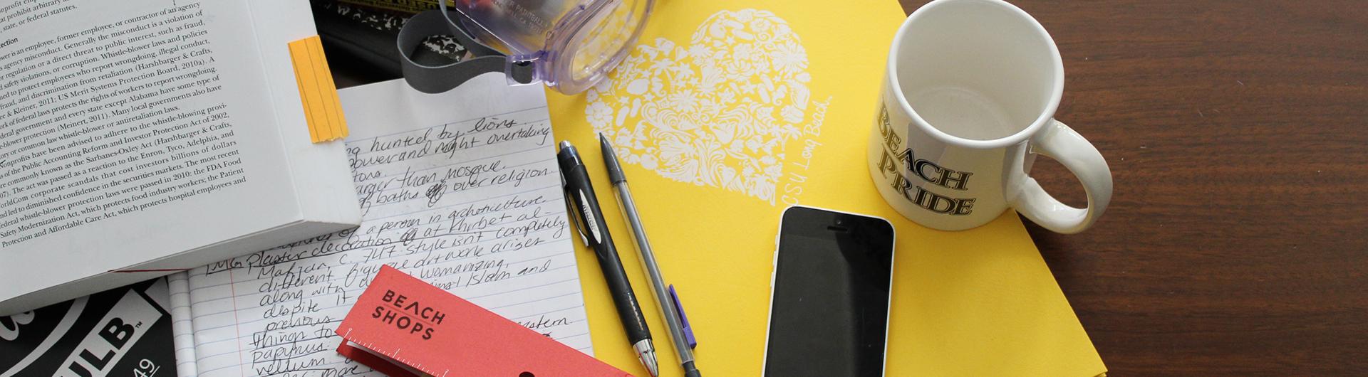 School supplies littered on a table with a beach pride coffee mug also. 